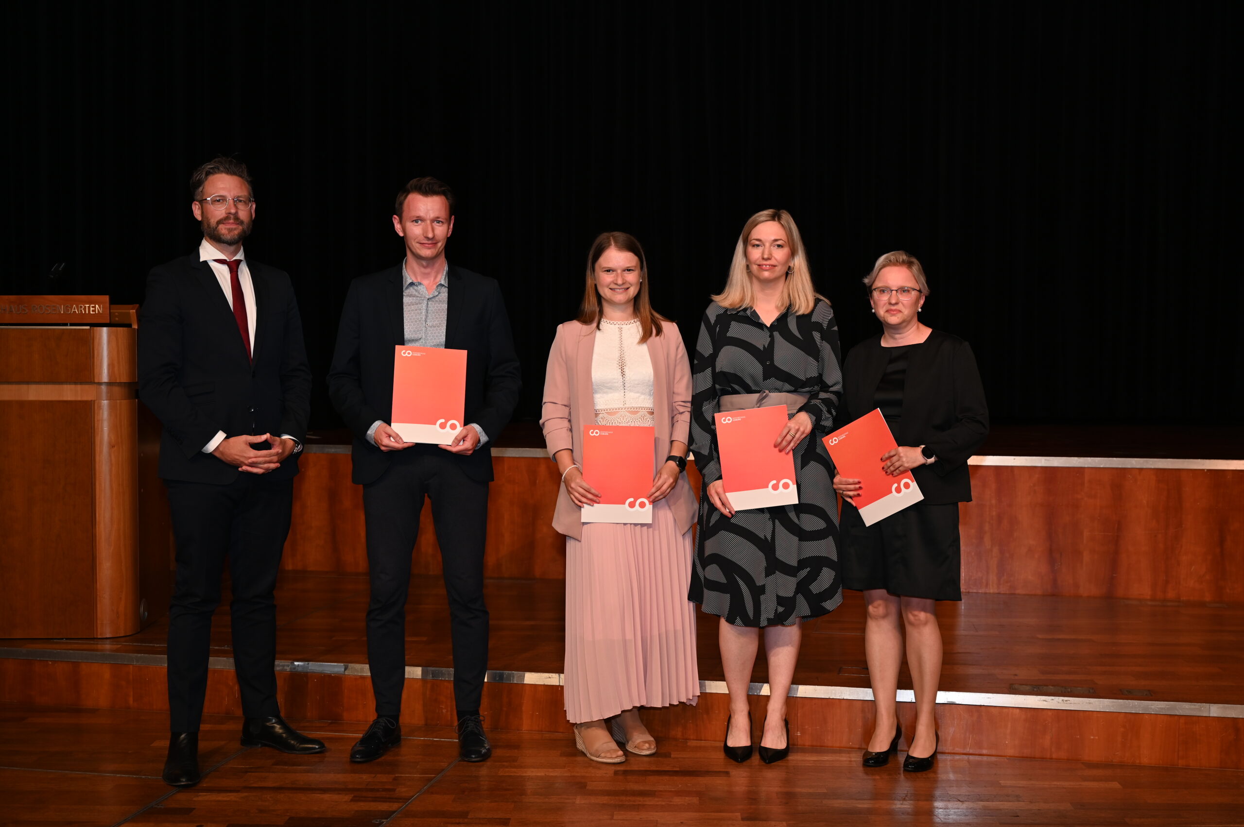 Fünf Personen stehen auf einer Bühne der Hochschule Coburg. Sie halten stolz ihre Zertifikate in den Händen und sind in formelle Kleidung gekleidet. Auf der linken Seite befindet sich ein Podium, vor einem Hintergrund dunkler Vorhänge, was den Eindruck einer angesehenen Preisverleihung erweckt.