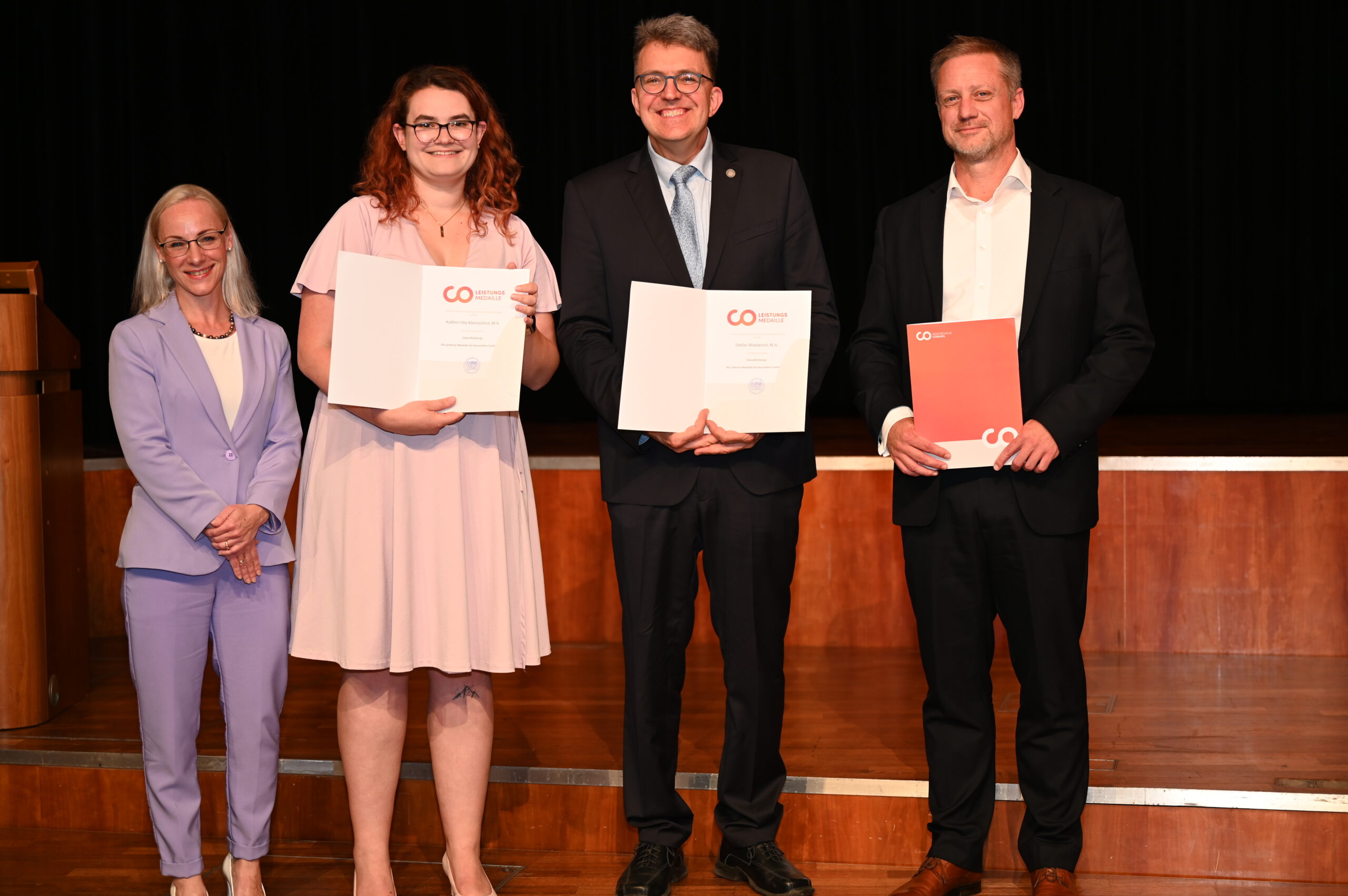 Vier formell gekleidete Personen stehen auf einer Bühne der Hochschule Coburg und halten Zertifikate in den Händen. Die beiden in der Mitte zeigen ihre Zertifikate offen, während die Person rechts ihr Zertifikat in einer roten Mappe hält. Im Hintergrund ist ein dunkler Vorhang.