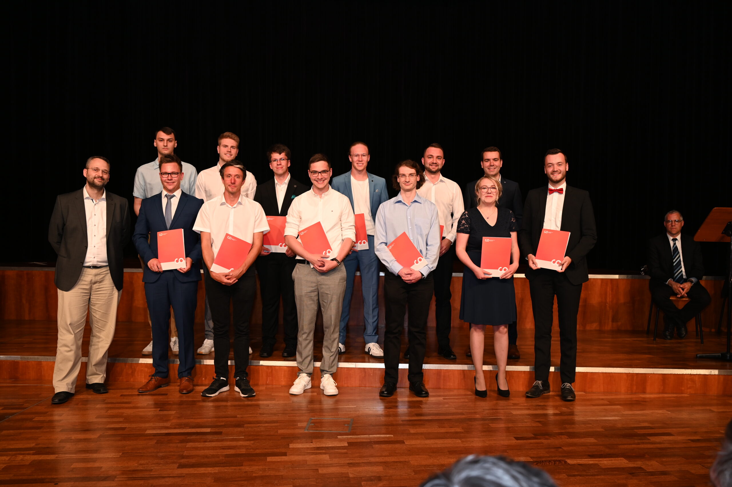 Eine Gruppe von Menschen steht auf einer Holzbühne der Hochschule Coburg und hält rote Mappen in der Hand. Sie sind in formelle Kleidung gekleidet. Hinter ihnen ist ein schwarzer Vorhang. Alle lächeln, und manche tragen Brillen und Fliegen.