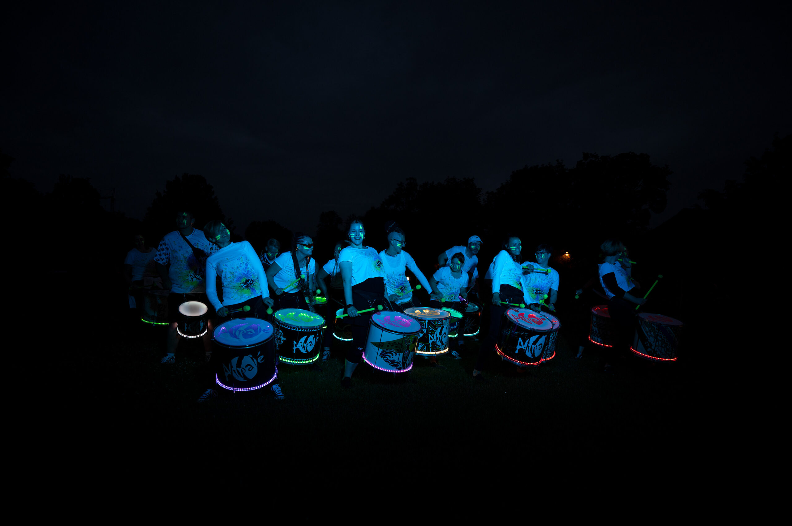 Eine Gruppe von Trommlern tritt nachts auf, beleuchtet durch leuchtende Neonlichter an ihren Trommeln und ihrer Kleidung. Ihre leuchtenden Farben heben sich vom dunklen Hintergrund ab und erzeugen eine lebendige und energiegeladene Szene, die an ein Festival der Hochschule Coburg erinnert.