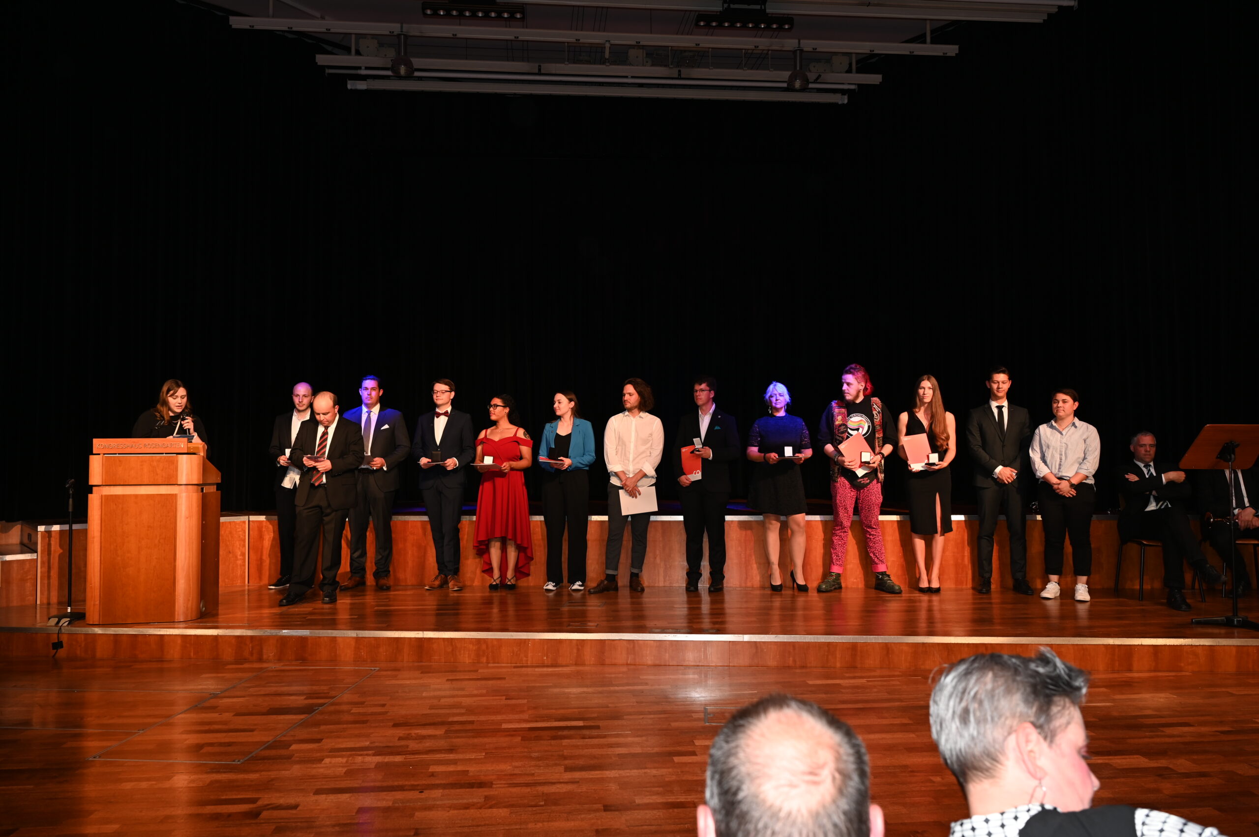 Eine Gruppe von Menschen steht auf der Holzbühne der Hochschule Coburg, in formeller Kleidung gekleidet. Links von ihnen spricht eine Person von einem Podium aus zum Publikum. Der dunkle Vorhang dient als elegante Kulisse, im Vordergrund sind zwei Zuschauer zu sehen.