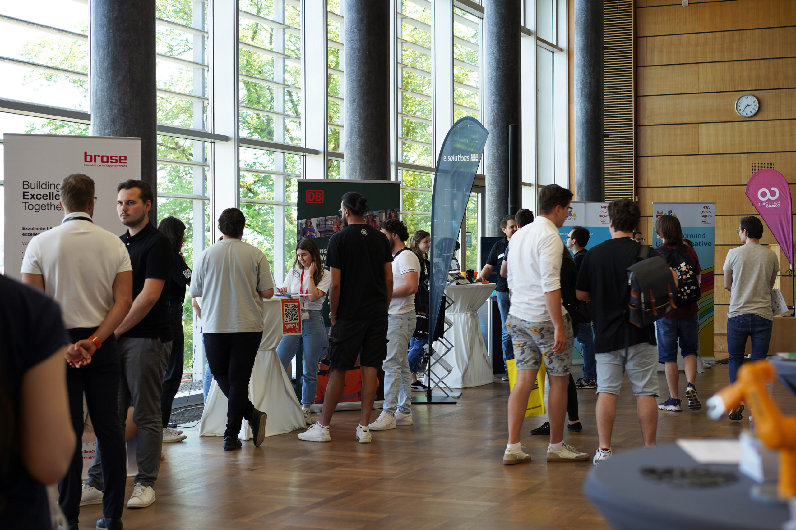 Menschen besuchen eine Jobmesse in einem großen Raum mit hohen Fenstern, die von der Hochschule Coburg veranstaltet wird. Es sind verschiedene Stände von Unternehmen aufgebaut und Besucher kommen mit Vertretern ins Gespräch. Auf Schildern und Bannern sind Firmenlogos und -informationen zu sehen, was zu einer geschäftigen Atmosphäre der Karrieremöglichkeiten beiträgt.