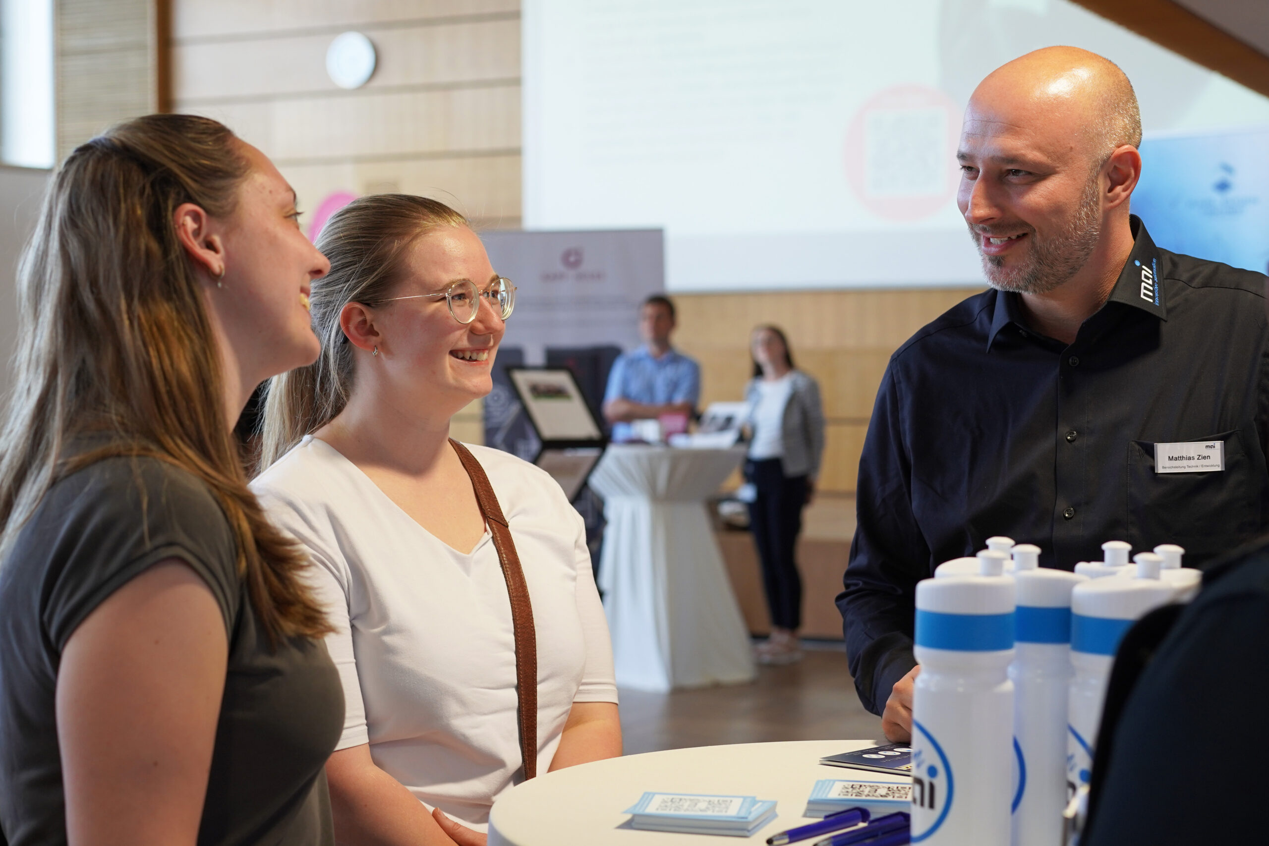 Drei Personen stehen an einem Tisch und unterhalten sich mit Werbewasserflaschen der Hochschule Coburg. Zwei Frauen stehen einem kahlköpfigen Mann in einem dunklen Hemd mit einem Namensschild gegenüber. Im Hintergrund ist eine Veranstaltungsumgebung mit geschäftigem Treiben, Präsentationen und Informationstafeln zu sehen.