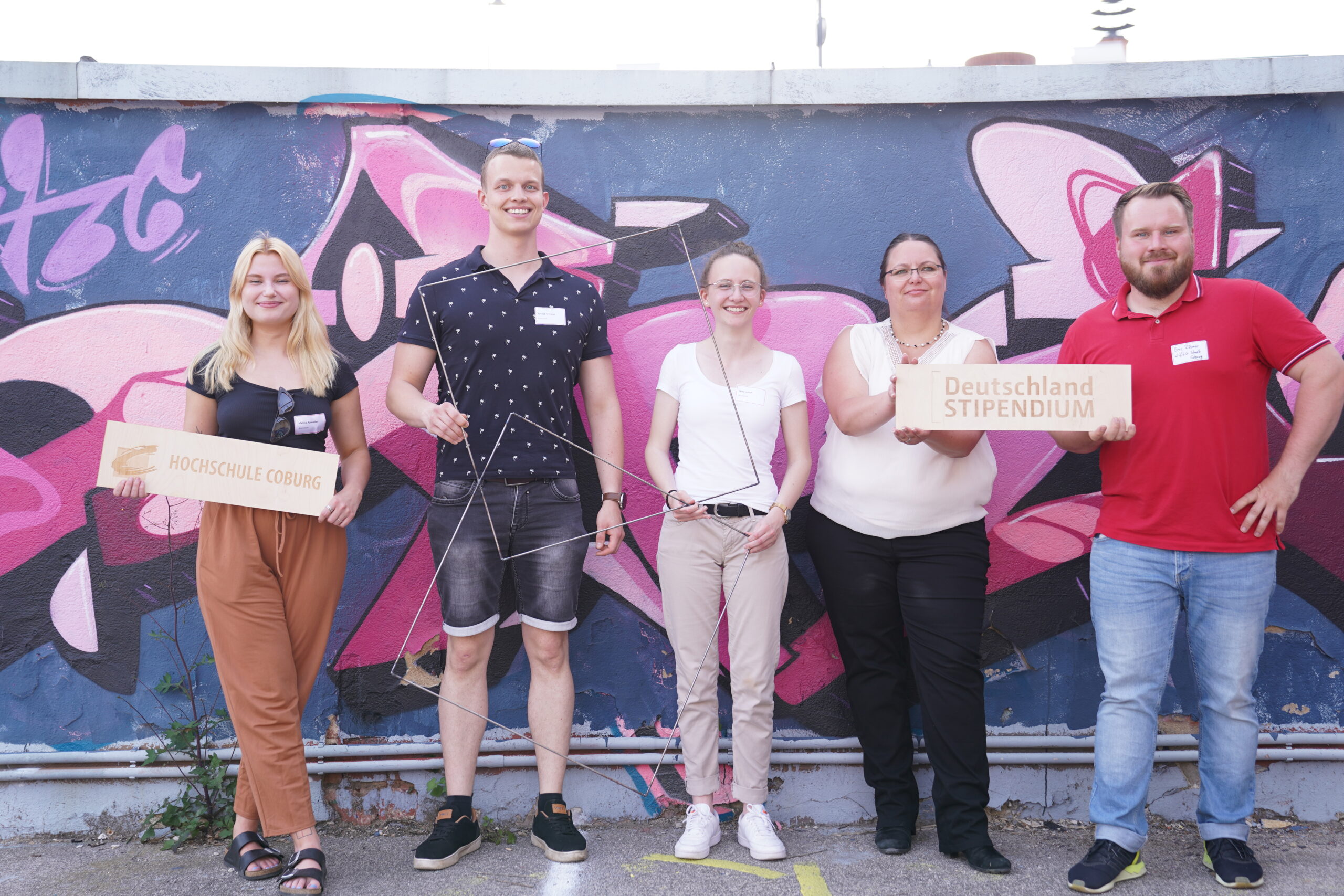 Fünf Personen stehen vor einer farbenfrohen Graffitiwand und halten stolz Schilder mit den Aufschriften „Hochschule Coburg“ und „Deutschlandstipendium“ hoch. Vier von ihnen strahlen vor Freude, während einer einen neutralen Gesichtsausdruck behält und damit den Geist dieser lebhaften Veranstaltung im Freien verkörpert.