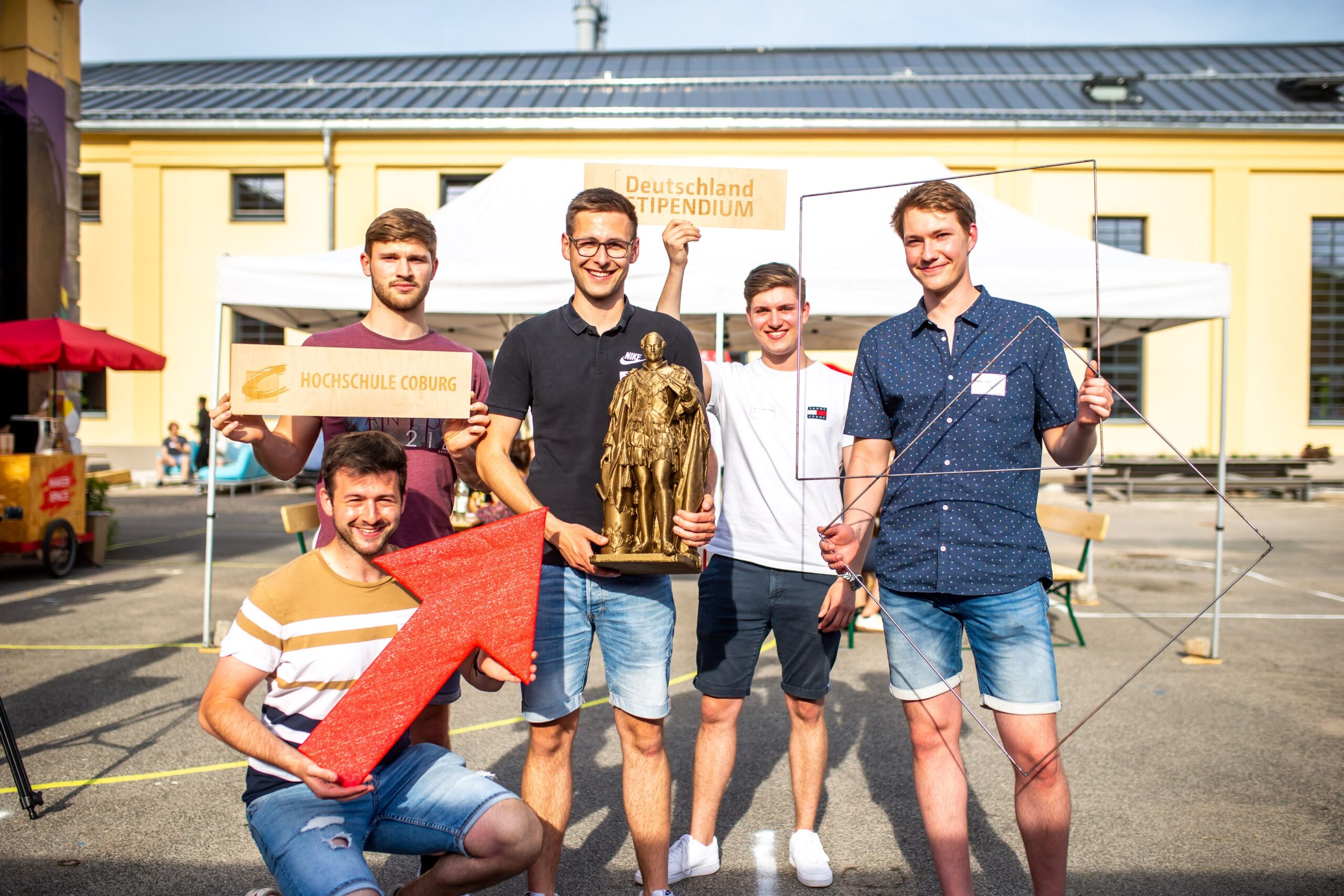 Fünf junge Männer stehen vor einem gelben Gebäude mit einem weißen Vordach und halten verschiedene Schilder hoch, darunter „Hochschule Coburg“ und „Deutschlandstipendium“, neben einem großen Pfeil und einem Drahtgittermodell der Eulerschen Formel.