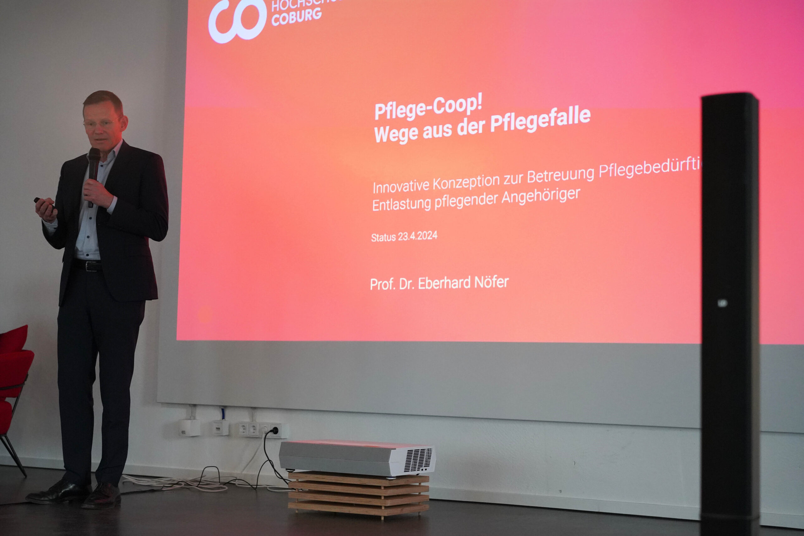 Ein Mann im Anzug präsentiert mit Mikrofon auf der Bühne der Hochschule Coburg. Hinter ihm läuft auf einer großen Leinwand ein Vortrag mit dem Titel „Pflege-Coop! Wege aus der Pflegefalle“ über ein innovatives Pflegekonzept von Prof. Dr. Eberhard Nofer vom 23.4.2024.