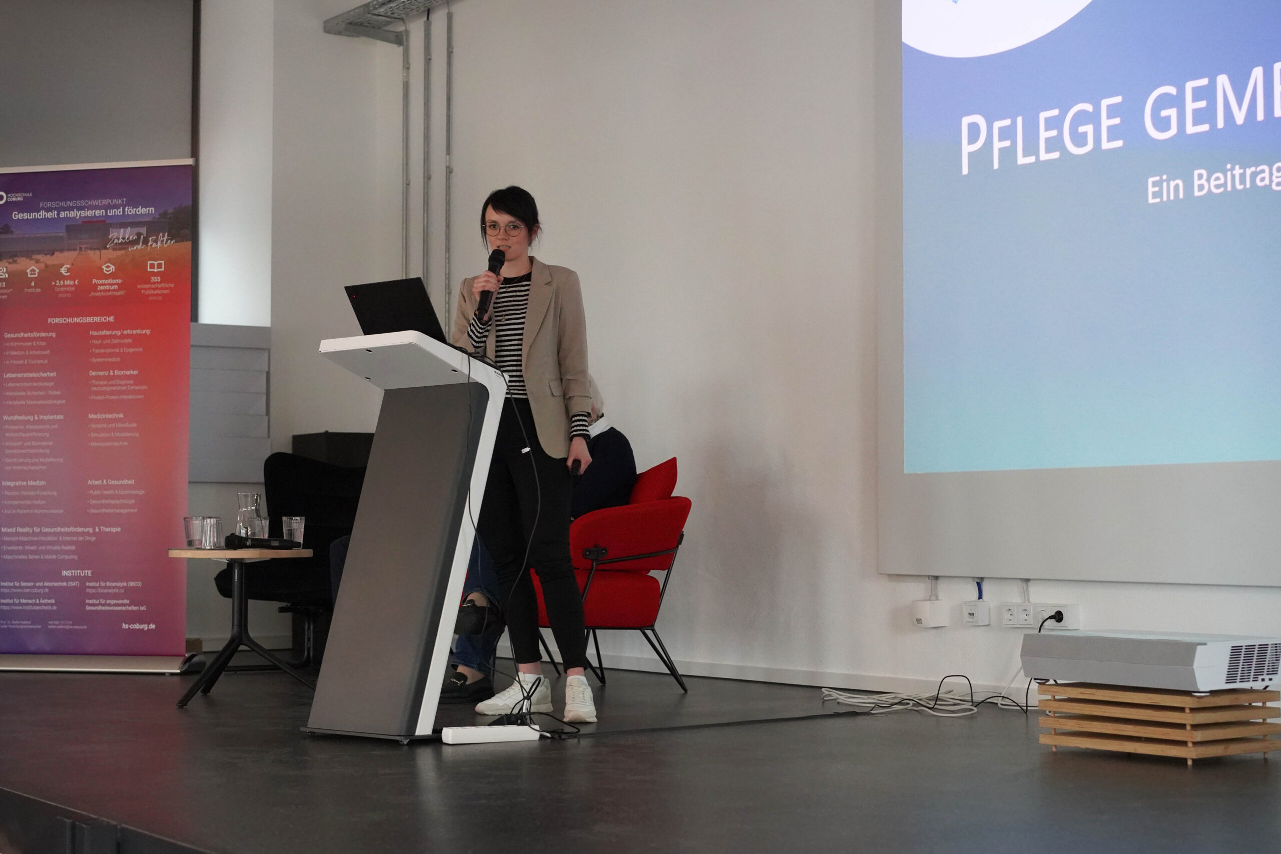 A person stands at a podium giving a presentation at Hochschule Coburg. They hold a microphone and are dressed in business casual attire, while the screen beside them displays a title in German. In the background, chairs and a banner further set the academic scene.