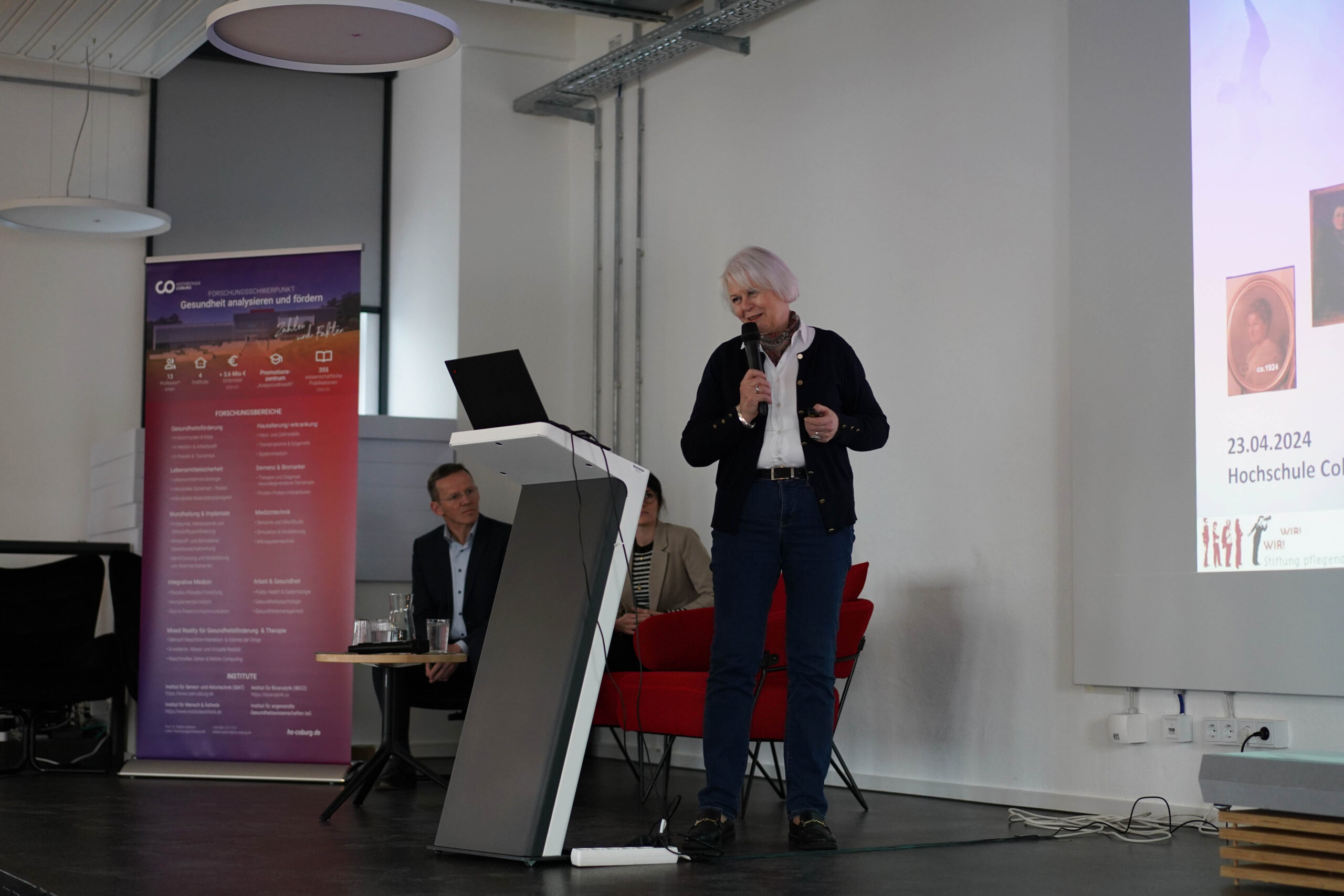 A person with white hair is delivering a speech at a Hochschule Coburg conference, using a microphone at the podium in front of a projected presentation slide. Two others are seated behind, attentively listening to the address.