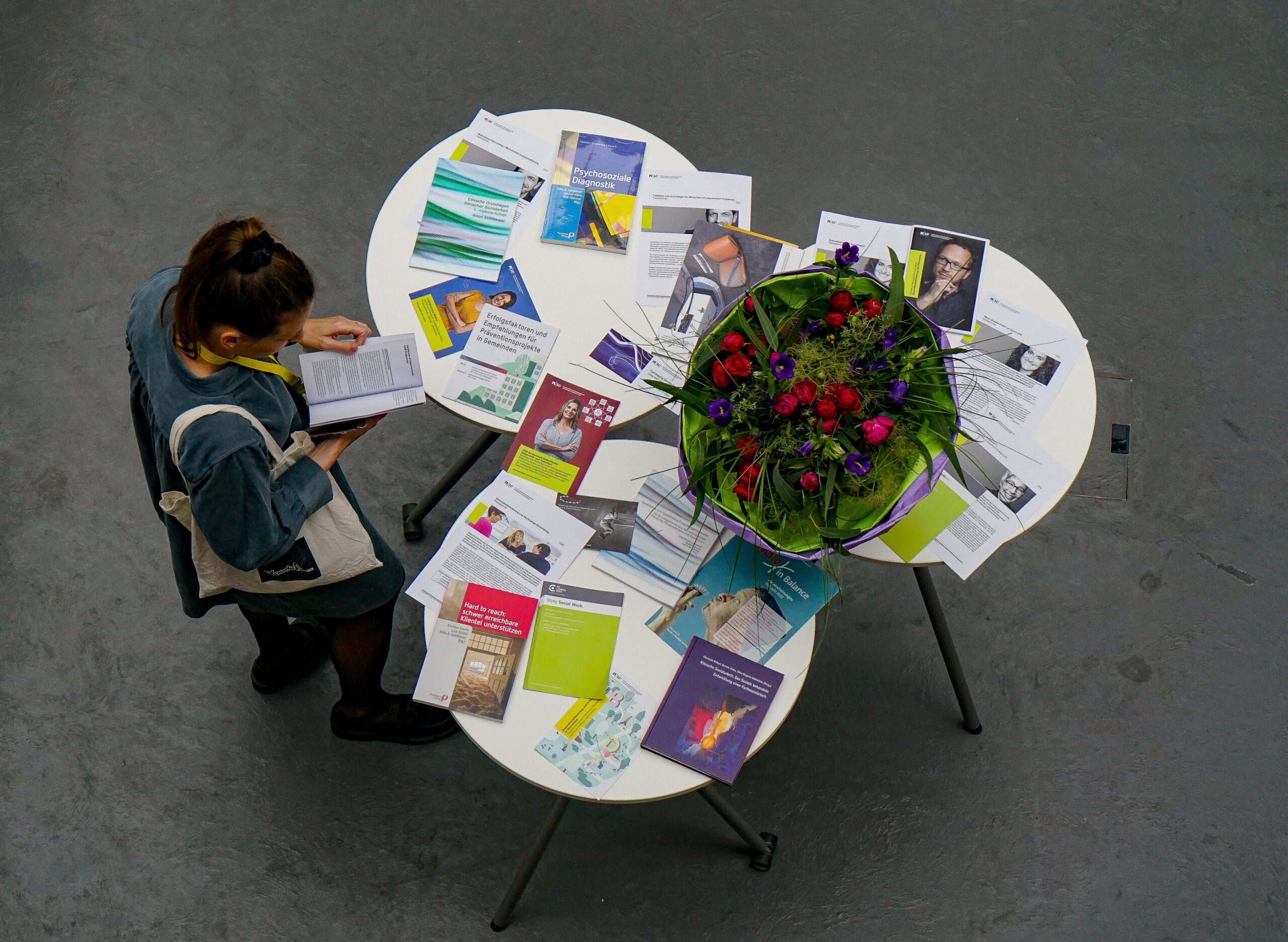 Eine Person steht an einem U-förmigen Tisch, der mit verschiedenen Büchern und Publikationen der Hochschule Coburg gefüllt ist. Auf einer Seite des Tisches befindet sich ein dekoratives Mittelstück mit leuchtenden Blumen. Die Person scheint eines der Dokumente zu lesen.