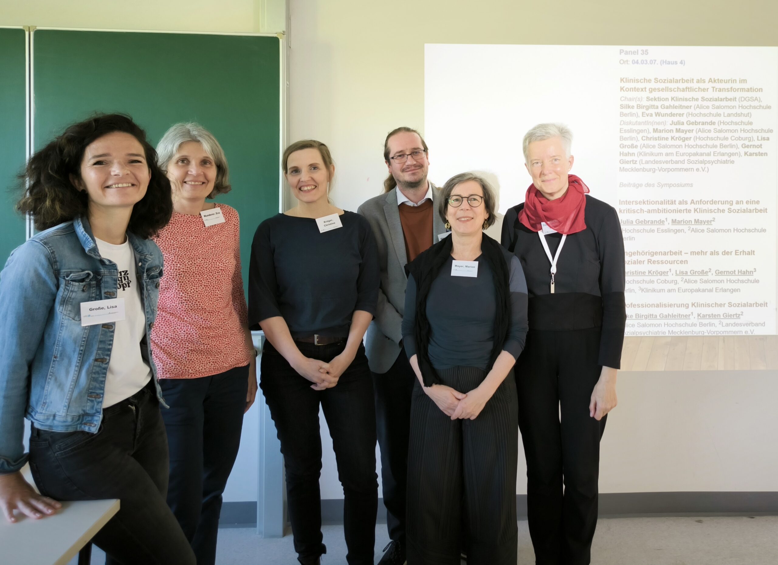 Eine Gruppe von sechs Erwachsenen steht in einem Hörsaal der Hochschule Coburg. Links steht eine grüne Tafel, an die Wand hinter ihnen wird eine Präsentationsfolie projiziert. Sie lächeln und tragen Konferenz-Namensschilder.