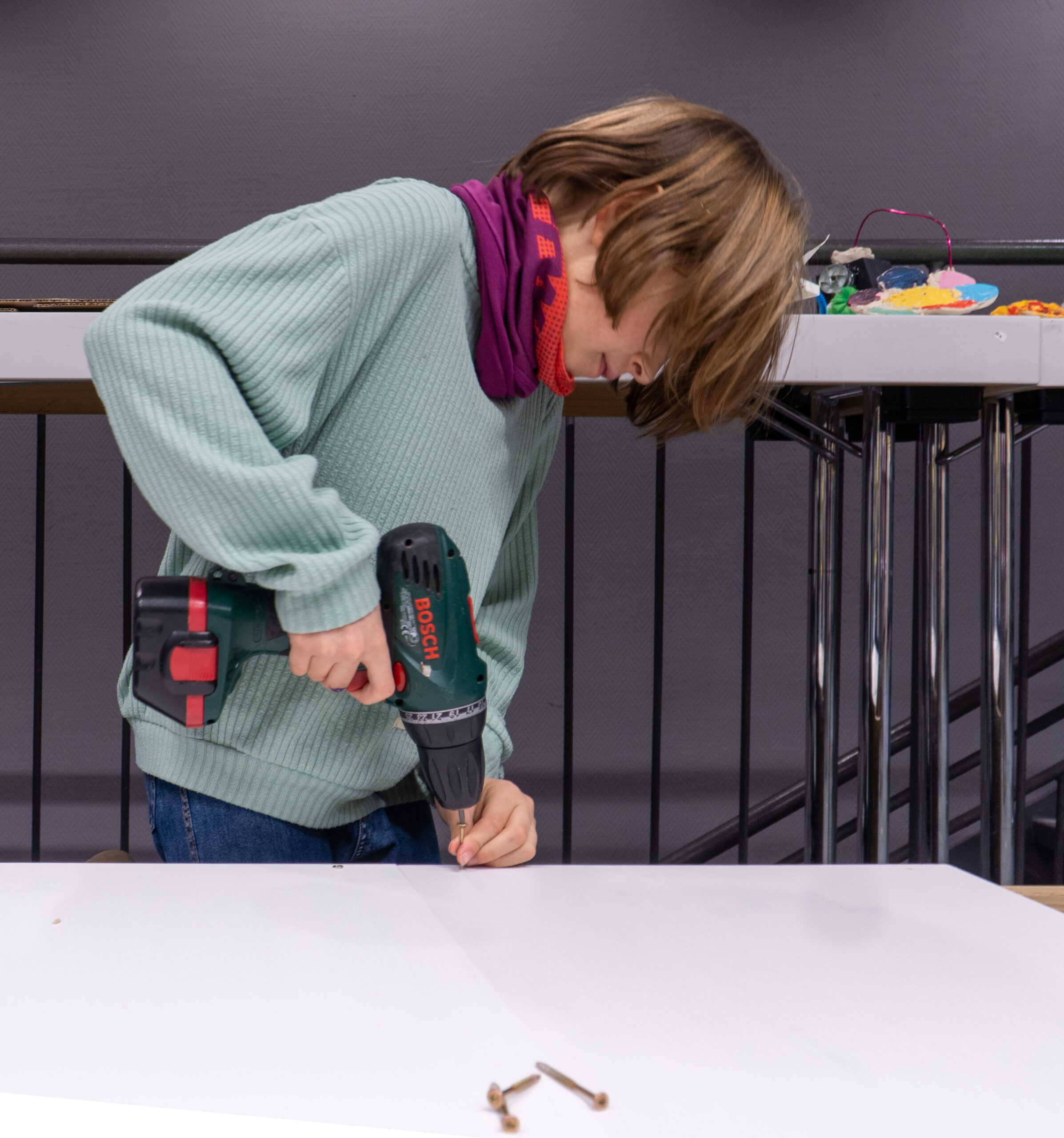 Eine Person in grünem Pullover und lila Schal schraubt mit einer Akku-Bohrmaschine ein Stück Holz fest. Vielleicht verfeinert sie ihre Fähigkeiten für ein Projekt an der Hochschule Coburg. Zwei Schrauben liegen auf dem weißen Tisch, vor dem Hintergrund eines schwach beleuchteten Raums mit schwarzem Geländer und einer grauen Wand.