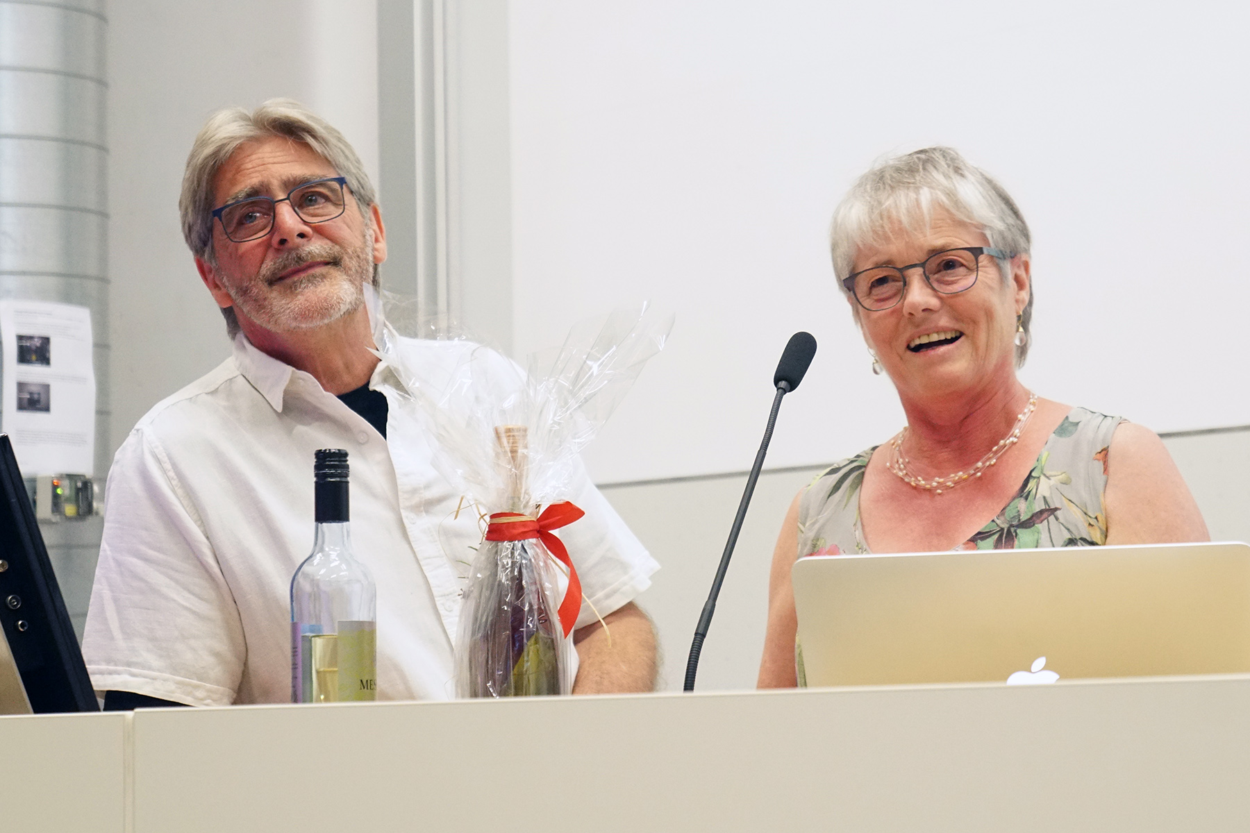 Ein älterer Mann und eine ältere Frau stehen lächelnd zusammen auf einem Podium der Hochschule Coburg. Der Mann hält ein verpacktes Geschenk und eine Flasche Wein in der Hand, während die Frau ins Mikrofon spricht. Ein Laptop und eine weitere Flasche stehen auf dem Podium und markieren einen besonderen Moment an dieser angesehenen Institution.