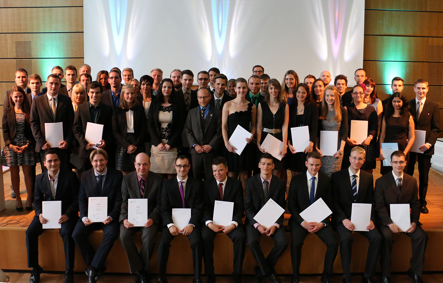 Eine große Gruppe von Menschen in formeller Kleidung posiert für ein Foto an der Hochschule Coburg. Viele halten Zertifikate in den Händen und stehen stolz auf einer Holzbühne, im Hintergrund eine weiße Leinwand und bunte Beleuchtung.