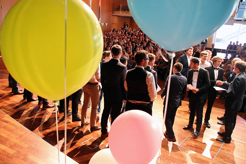 Eine große Gruppe formell gekleideter Menschen versammelt sich in einem Saal mit Holzboden in der Hochschule Coburg. Bunte Luftballons in Gelb, Rosa und Blau tragen zur festlichen Atmosphäre bei und deuten auf ein besonderes Ereignis oder eine Zeremonie hin, an der viele teilnehmen.