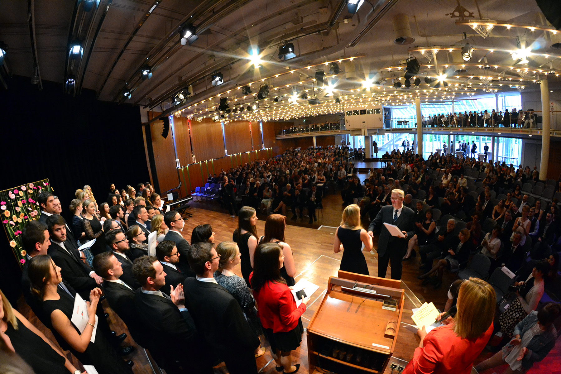 Ein großer Chor der Hochschule Coburg tritt in einem Konzertsaal auf, der mit einem begeisterten Publikum gefüllt ist. Der Saal ist mit Lichterketten geschmückt, und die Menschen sitzen und stehen und genießen die lebendige und festliche Atmosphäre.