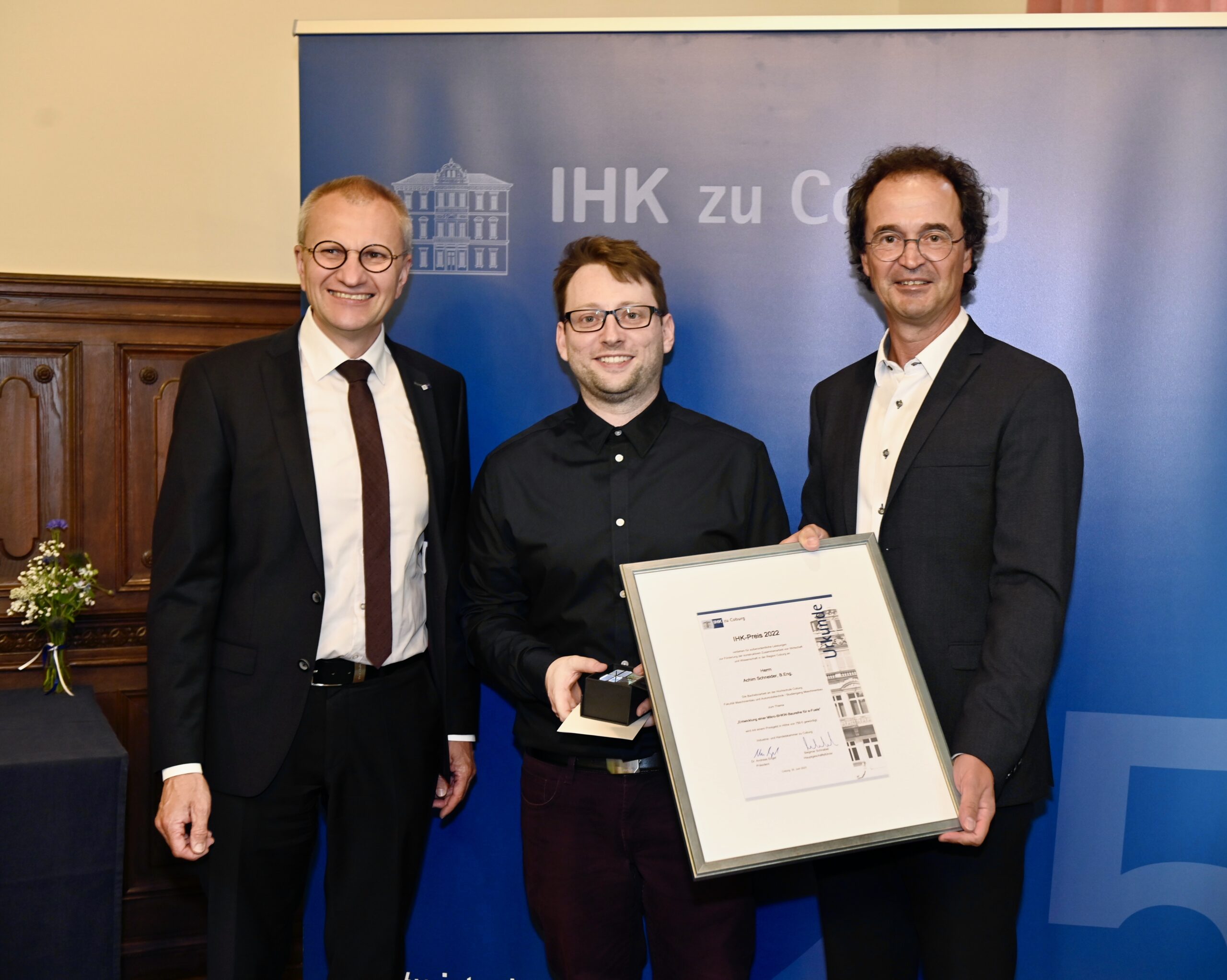 Drei Männer stehen zusammen und lächeln vor einem blauen Banner mit der Aufschrift „IHK zu Coburg“. Der Mann in der Mitte, der wahrscheinlich die Hochschule Coburg vertritt, hält ein gerahmtes Zertifikat und eine kleine Trophäe in den Händen. Sie tragen formelle Kleidung und auf einem Tisch links steht ein Blumenstrauß.