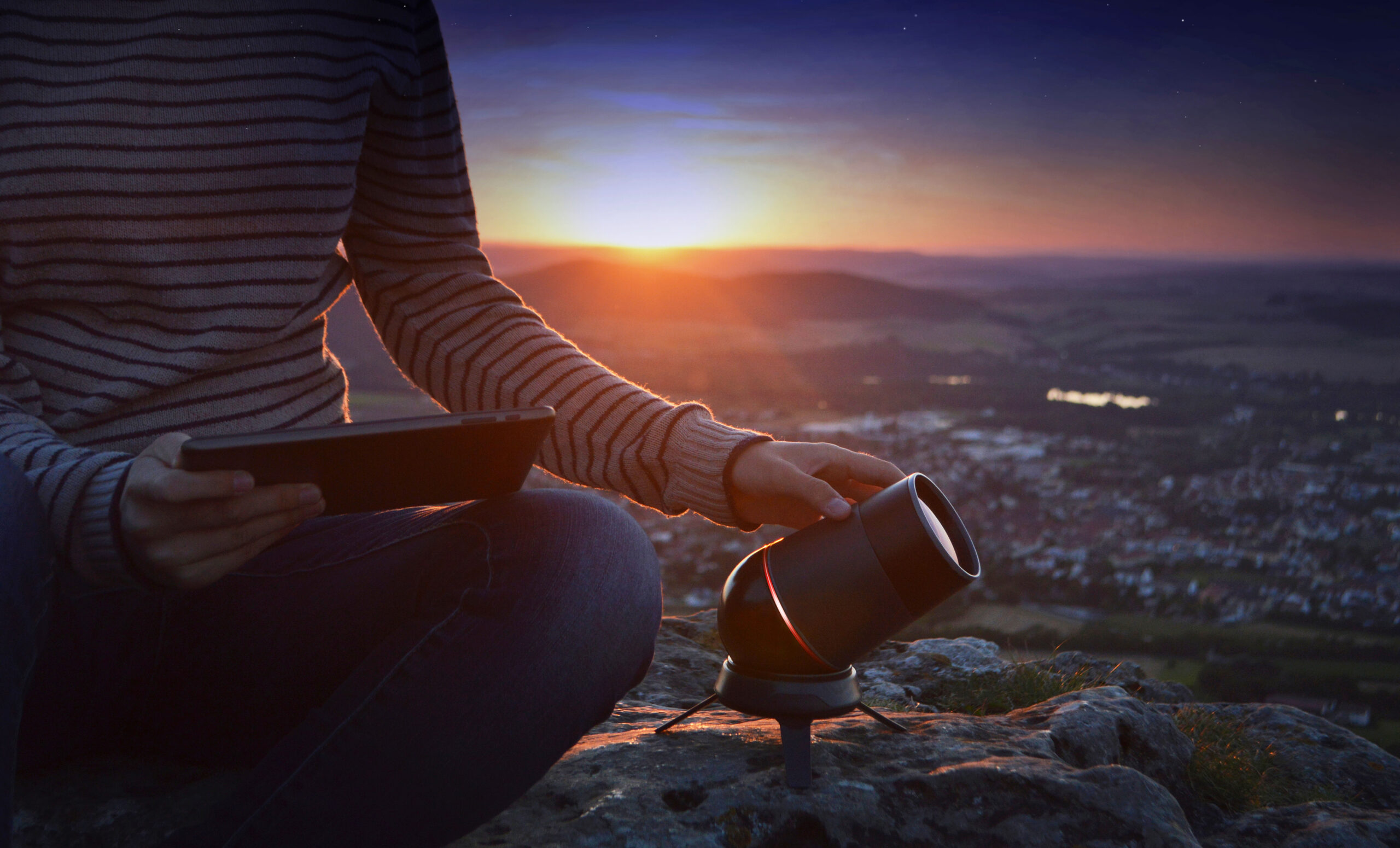 Eine Person sitzt bei Sonnenuntergang auf einem felsigen Hügel, hält ein Tablet in der Hand und justiert ein kleines Teleskop, ganz wie ein Astronomiestudent der Hochschule Coburg. Die Landschaft unter ihnen erstreckt sich bis zum Horizont, getaucht in das sanfte Licht der untergehenden Sonne.