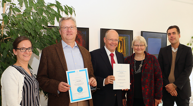 Fünf Personen der Hochschule Coburg stehen drinnen, lächeln und halten Zertifikate in den Händen. Sie tragen formelle Kleidung, im Hintergrund ist eine große Pflanze zu sehen. An der Wand hängen gerahmte Kunstwerke.