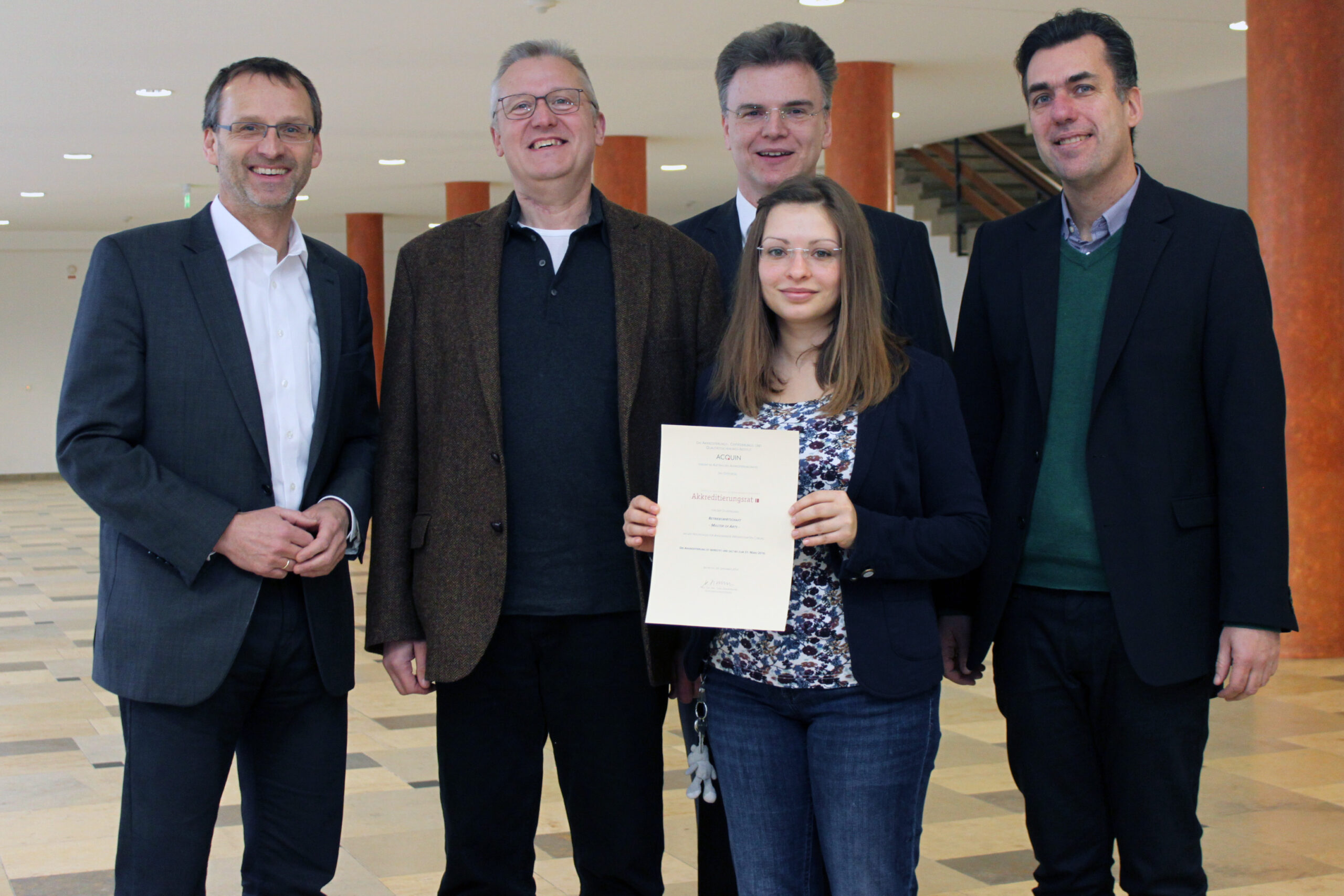 Eine Frau hält stolz ein Zertifikat der Hochschule Coburg in den Händen, umgeben von vier Männern in Business-Kleidung. Sie stehen drinnen vor einem neutralen Hintergrund und orangefarbenen Säulen und lächeln alle herzlich in die Kamera.