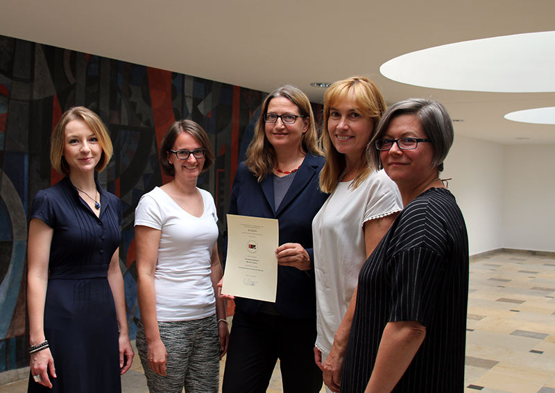 Fünf Frauen stehen in einem Raum der Hochschule Coburg zusammen. Eine hält ein Zertifikat in der Hand. Sie lächeln alle, im Hintergrund eine gemusterte Wand und darüber runde Deckenleuchten.