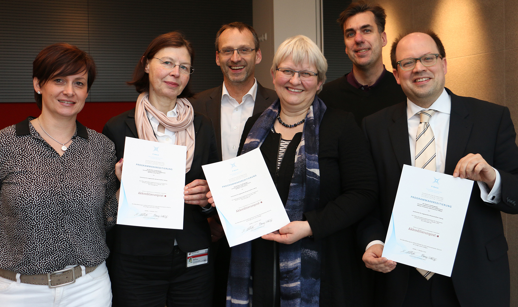 Eine Gruppe von sechs Personen der Hochschule Coburg lächelt in einem Innenbereich, drei halten stolz ihre Zertifikate in den Händen. Sie scheinen an einer Veranstaltung oder Zeremonie teilzunehmen, sind in Business-Kleidung gekleidet und stehen vor einem Hintergrund, der aus einer Mischung dunkler und heller Farben besteht.