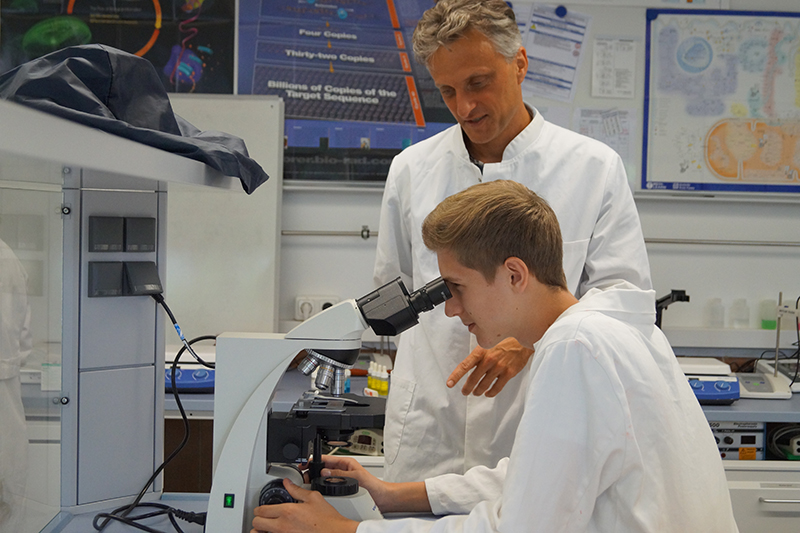 In einem mit wissenschaftlichen Postern geschmückten Labor an der Hochschule Coburg blickt ein junger Mann im Laborkittel durch ein Mikroskop, während ein älterer Mentor neben ihm steht und ihm Anleitung gibt.