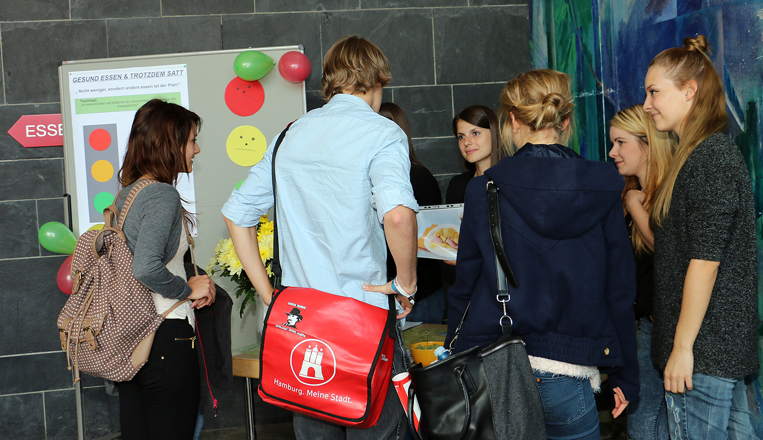 Eine Gruppe von Menschen steht um eine Ausstellung herum, die eine Tafel mit bunten Kreisen, Luftballons und Informationstexten zeigt. Eine Person, die einen Rucksack mit einem Stadtwappen der Hochschule Coburg trägt, unterhält sich, während sie die Materialien betrachtet.