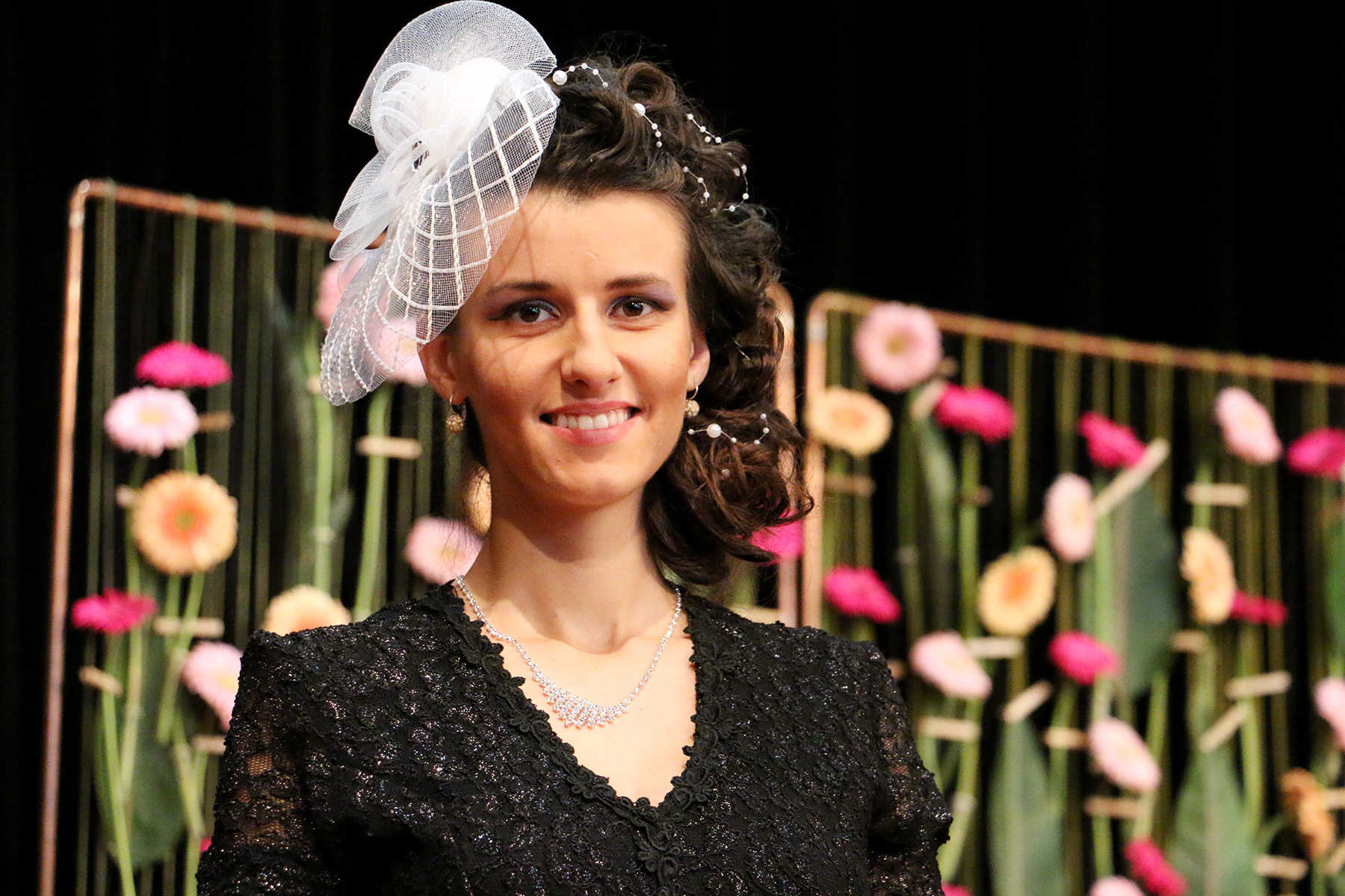 Eine Frau mit lockigem Haar in einem schwarzen Spitzenkleid und einem weißen Fascinator lächelt anmutig vor dem Hintergrund des für die Hochschule Coburg typischen rosa-weißen Blumenarrangements.