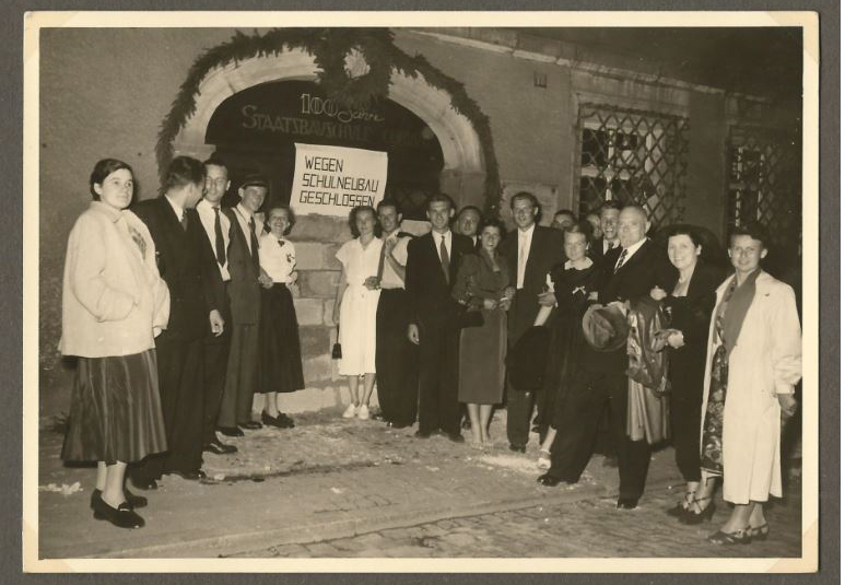 Eine Gruppe von Menschen steht vor einem Gebäude, das mit einem Banner geschmückt ist, auf dem „100 Jahre Staatsbrauhaus“ gefeiert wird. Auf dem Banner wird auch eine Schließung erwähnt. Einige sind in Kleidung aus der Mitte des 20. Jahrhunderts gekleidet und lächeln in die Kamera. Möglicherweise handelt es sich um Alumni der Hochschule Coburg, die sich treffen, um das Erbe zu ehren.