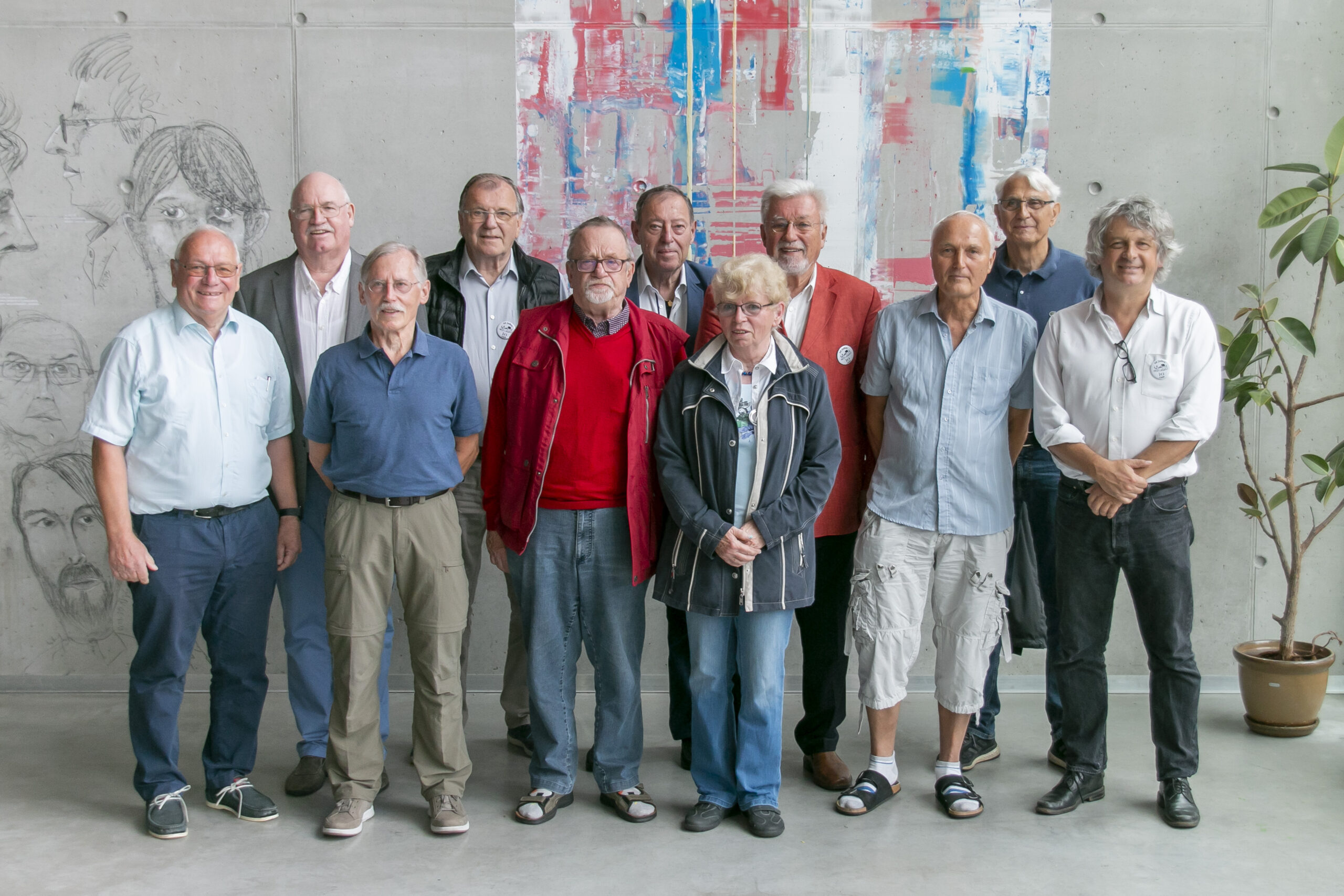 Eine Gruppe von elf älteren Erwachsenen, allesamt Männer, steht lächelnd in einer Reihe an der Hochschule Coburg. Sie befinden sich in einem modernen Ambiente mit grauen Wänden, an denen künstlerische Zeichnungen und ein farbenfrohes abstraktes Gemälde hängen. Auf der rechten Seite steht eine Topfpflanze.