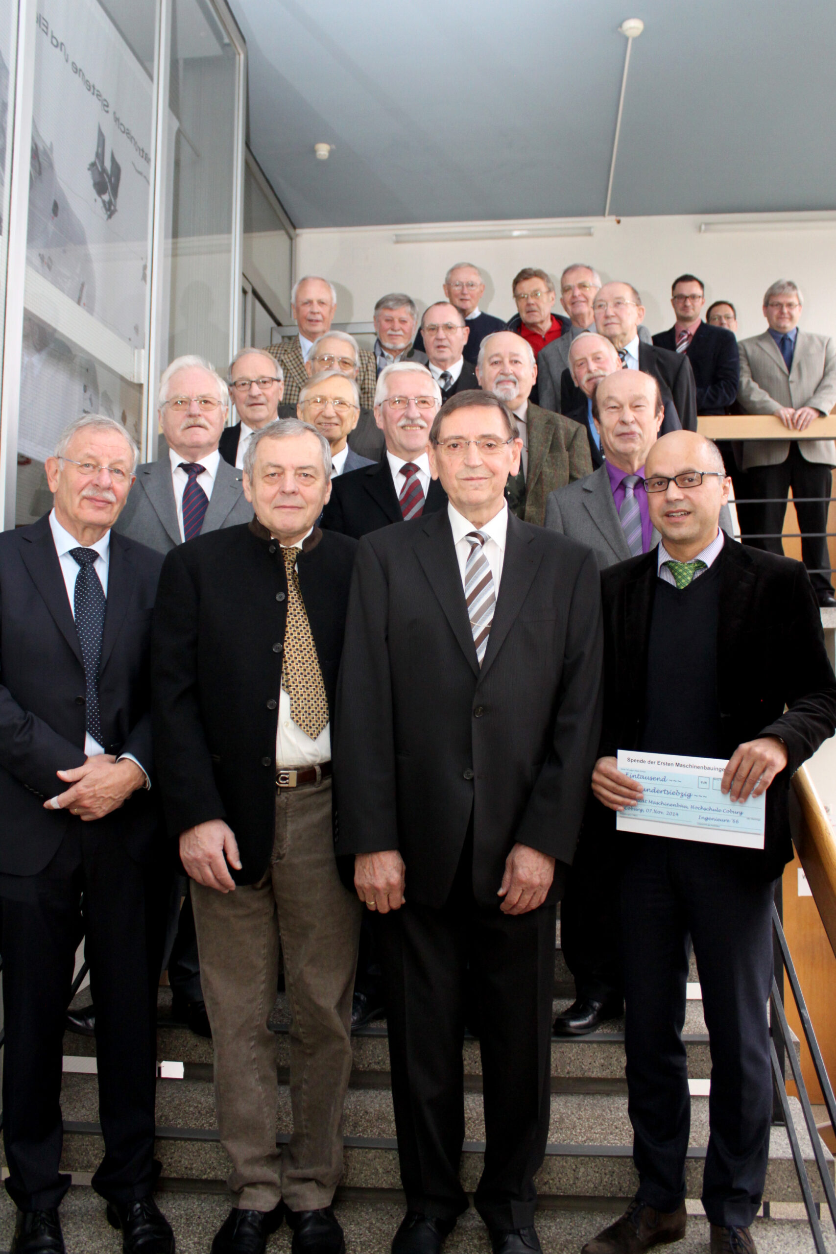 Eine Gruppe älterer Männer in Anzug und Krawatte steht zusammen auf einer Treppe der Hochschule Coburg. In der ersten Reihe steht ein Mann, der einen großen Scheck hält. Die Szene scheint sich in einem Innenbereich zu befinden, möglicherweise in einem Büro- oder Institutsgebäude.