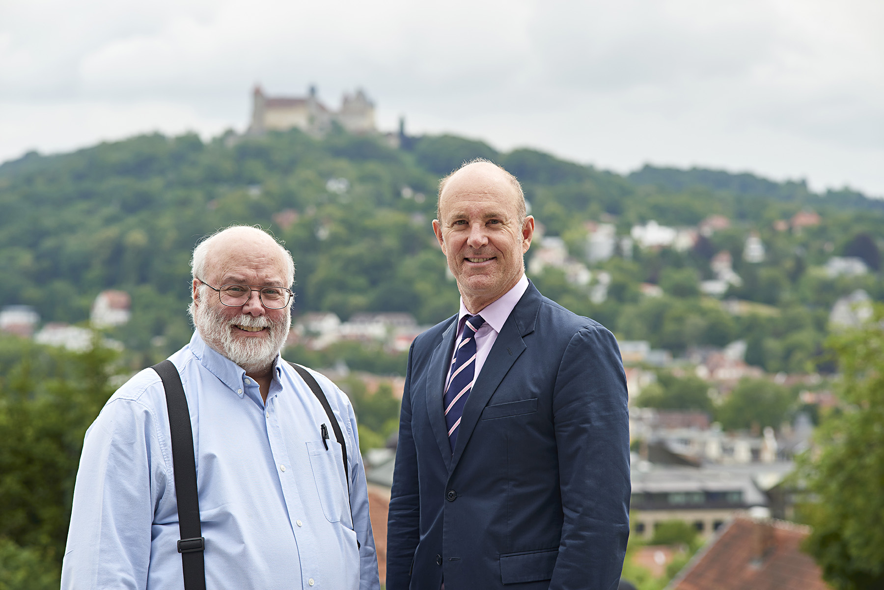 Zwei ältere Männer stehen draußen vor einem malerischen Hügel und einer Burg im Hintergrund und verkörpern den Charme der Hochschule Coburg. Einer trägt ein hellblaues Hemd mit Hosenträgern, der andere einen Anzug mit gestreifter Krawatte. Beide lächeln herzlich in die Kamera.