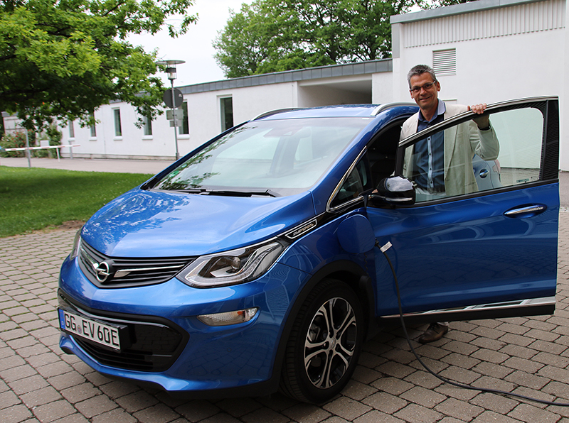 Eine Person steht neben einem blauen Elektroauto in der Nähe der Hochschule Coburg und hält die Fahrertür offen. Das Auto steht auf einer gepflasterten Fläche und hat ein angeschlossenes Ladekabel. Im Hintergrund sind Bäume und weiße Gebäude zu sehen, die die pädagogische Atmosphäre ergänzen.