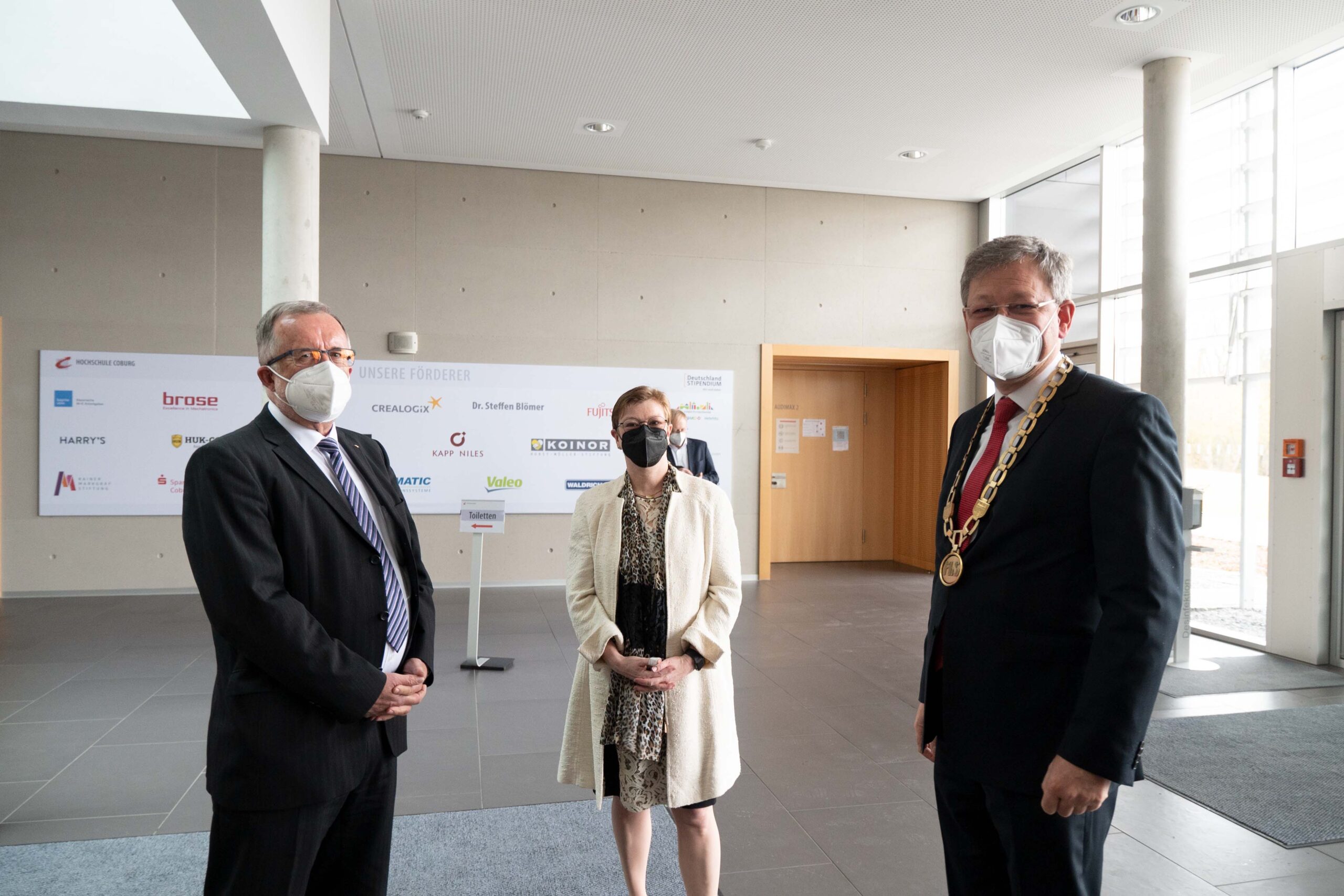 Drei Menschen mit Masken stehen in einem geräumigen Raum mit großen Fenstern. Ein Mann, geschmückt mit einer Dienstkette, deutet an, dass es sich um einen offiziellen Besuch der Hochschule Coburg handeln könnte. Im Hintergrund ist ein Schild mit Logos zu sehen, das der Versammlung einen Kontext verleiht.