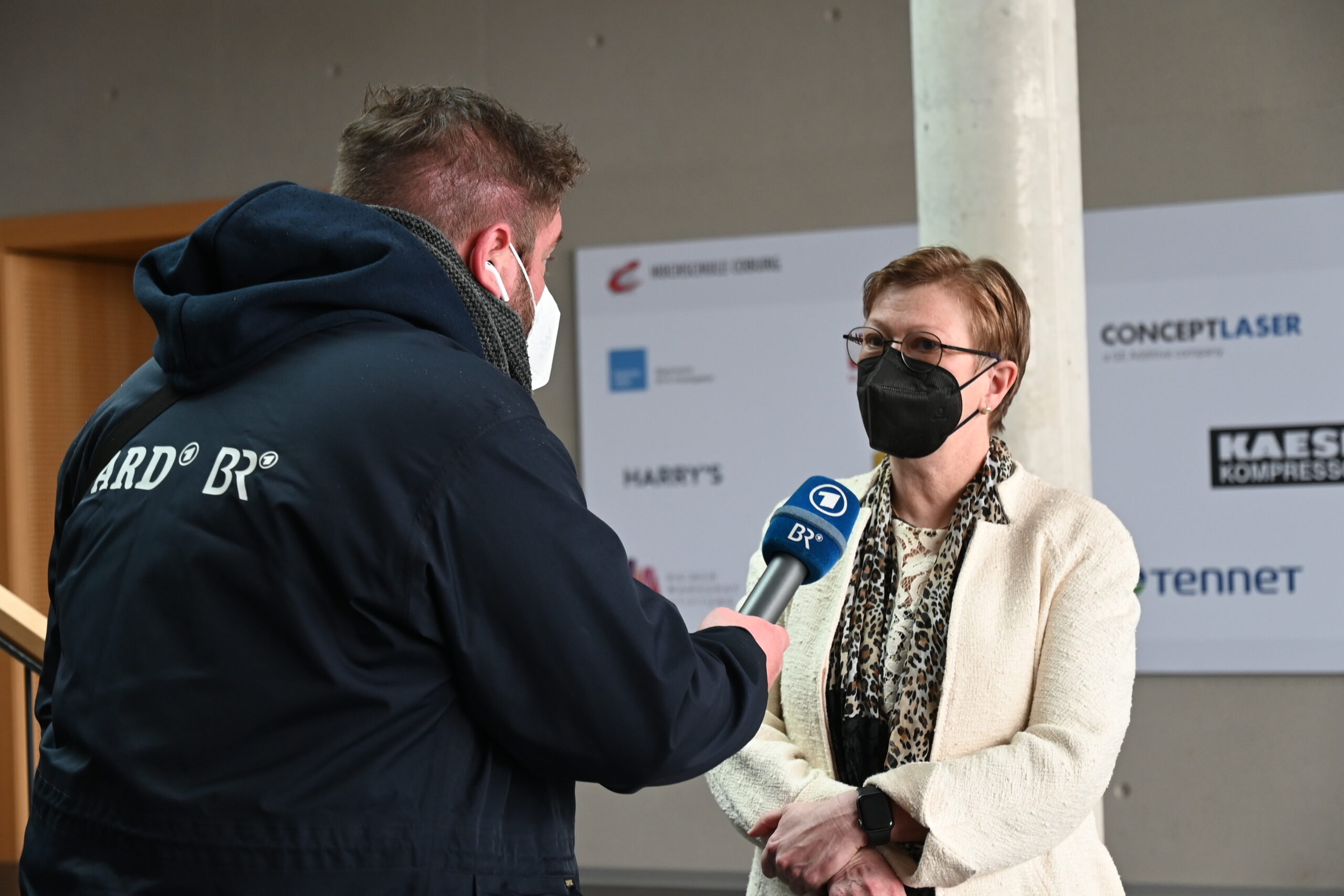 Eine Person mit einer schwarzen Maske und einem Mikrofon in der Hand interviewt eine Frau in einer beigen Jacke und einer schwarzen Maske, die vor einer Wand steht, die mit den Logos der Hochschule Coburg und anderer Unternehmen geschmückt ist.