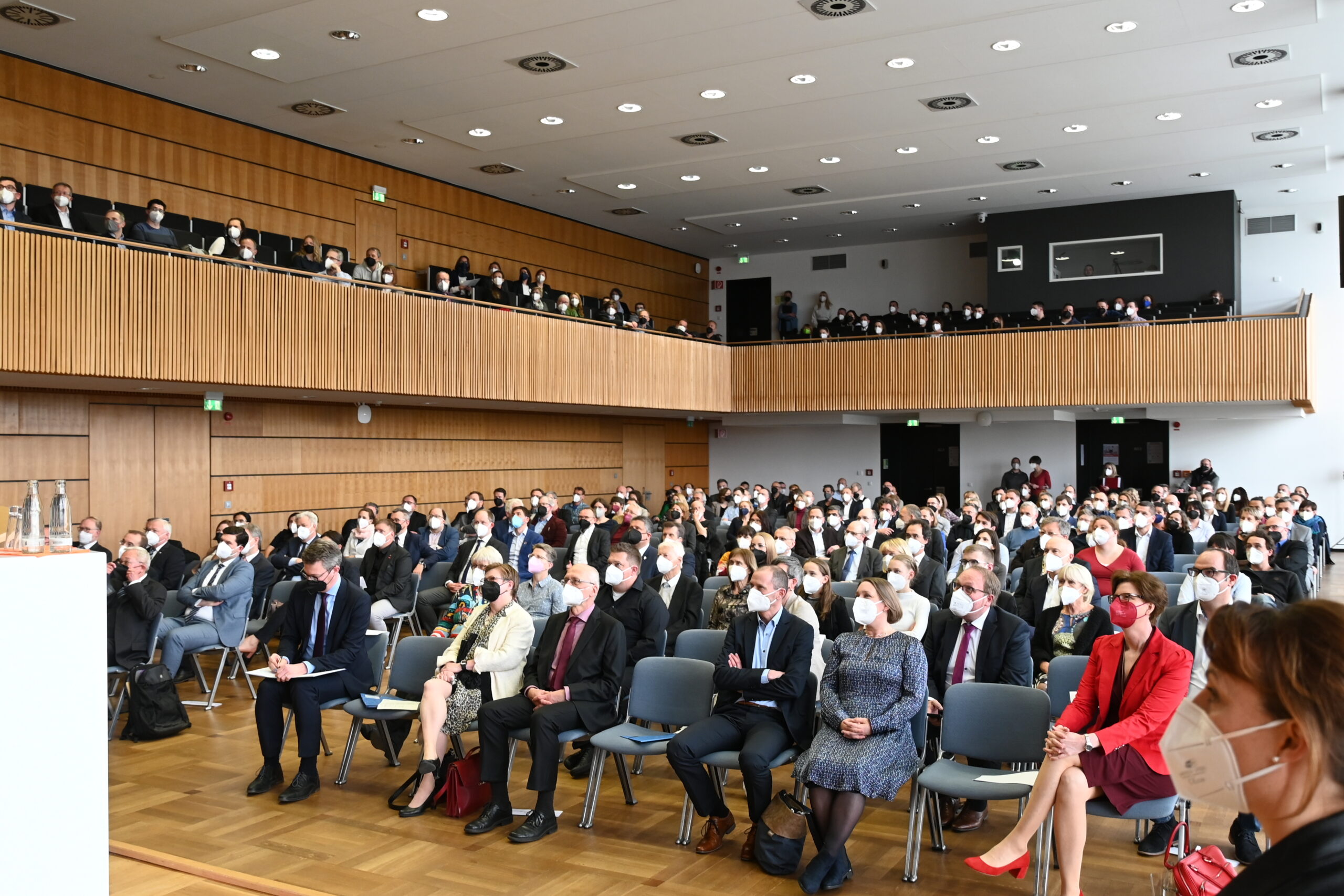 Eine große Gruppe von Menschen sitzt in einem modernen Konferenzraum mit Holzakzenten an der Hochschule Coburg. Einige Teilnehmer tragen Masken, einige andere stehen im Hintergrund und auf einem oberen Balkon und blicken aufmerksam nach vorne in den Raum.