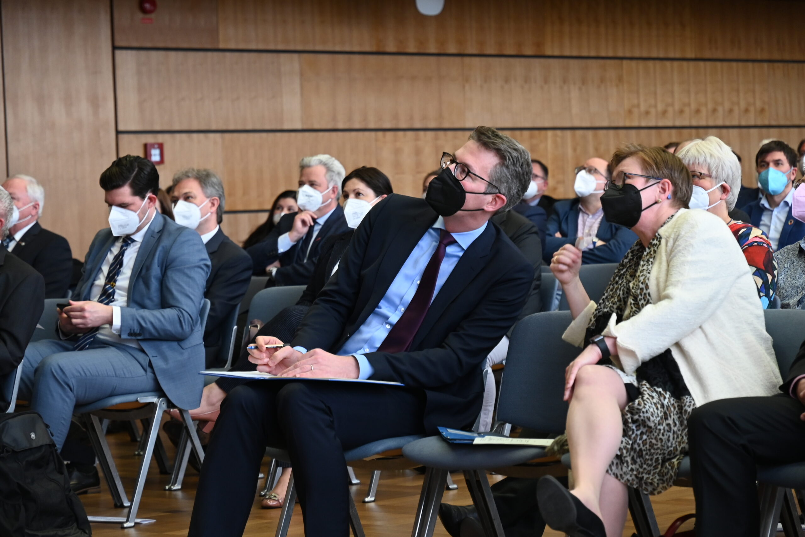 In einem Konferenzraum der Hochschule Coburg sitzt eine Gruppe maskierter Menschen und hört aufmerksam zu. Einige machen sich Notizen, im Vordergrund sind eine Frau und ein Mann einander leicht zugewandt und in ein Gespräch vertieft.