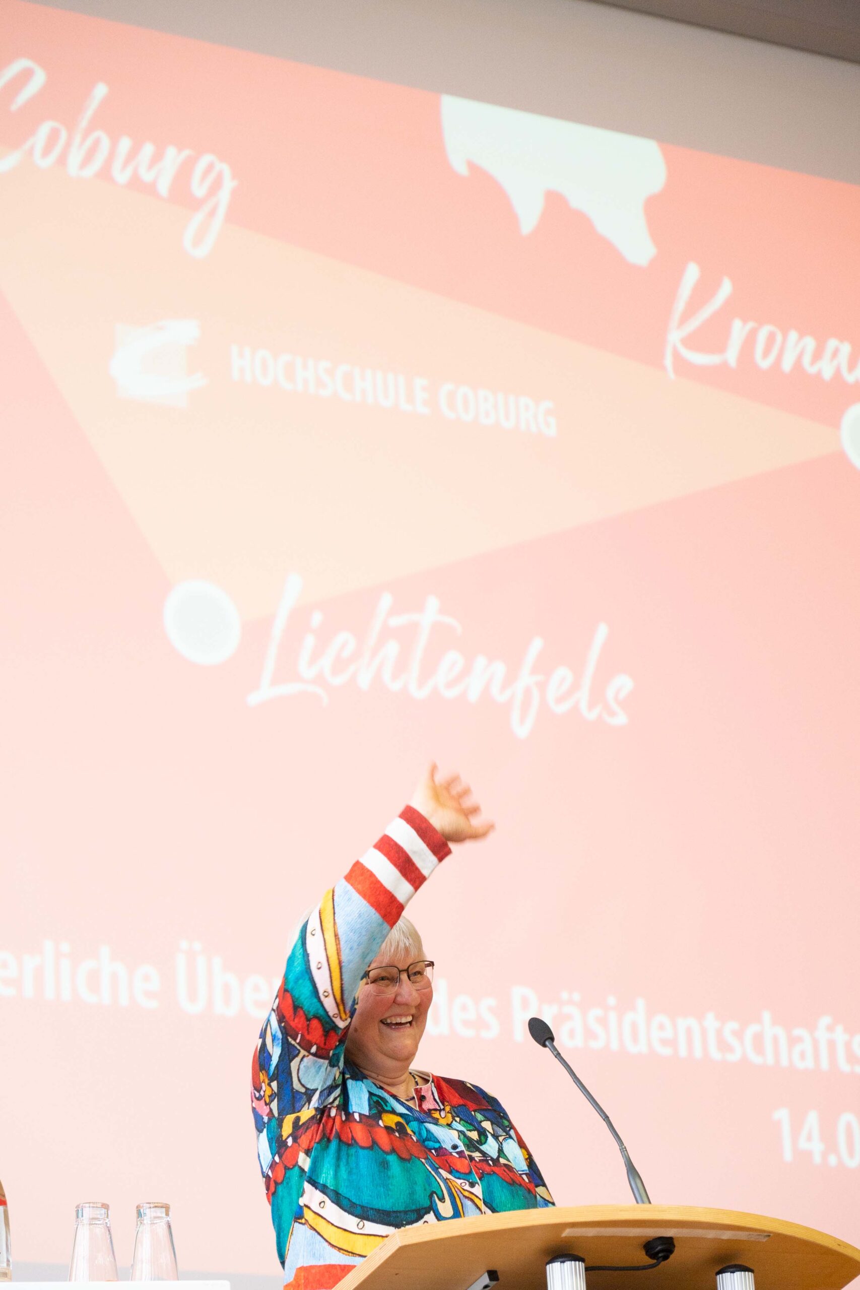 Eine lebhafte Person in einem bunten Oberteil steht selbstbewusst lächelt und winkt an einem Podium vor einem roten Hintergrund, der mit Texten und Logos der Hochschule Coburg geschmückt ist.