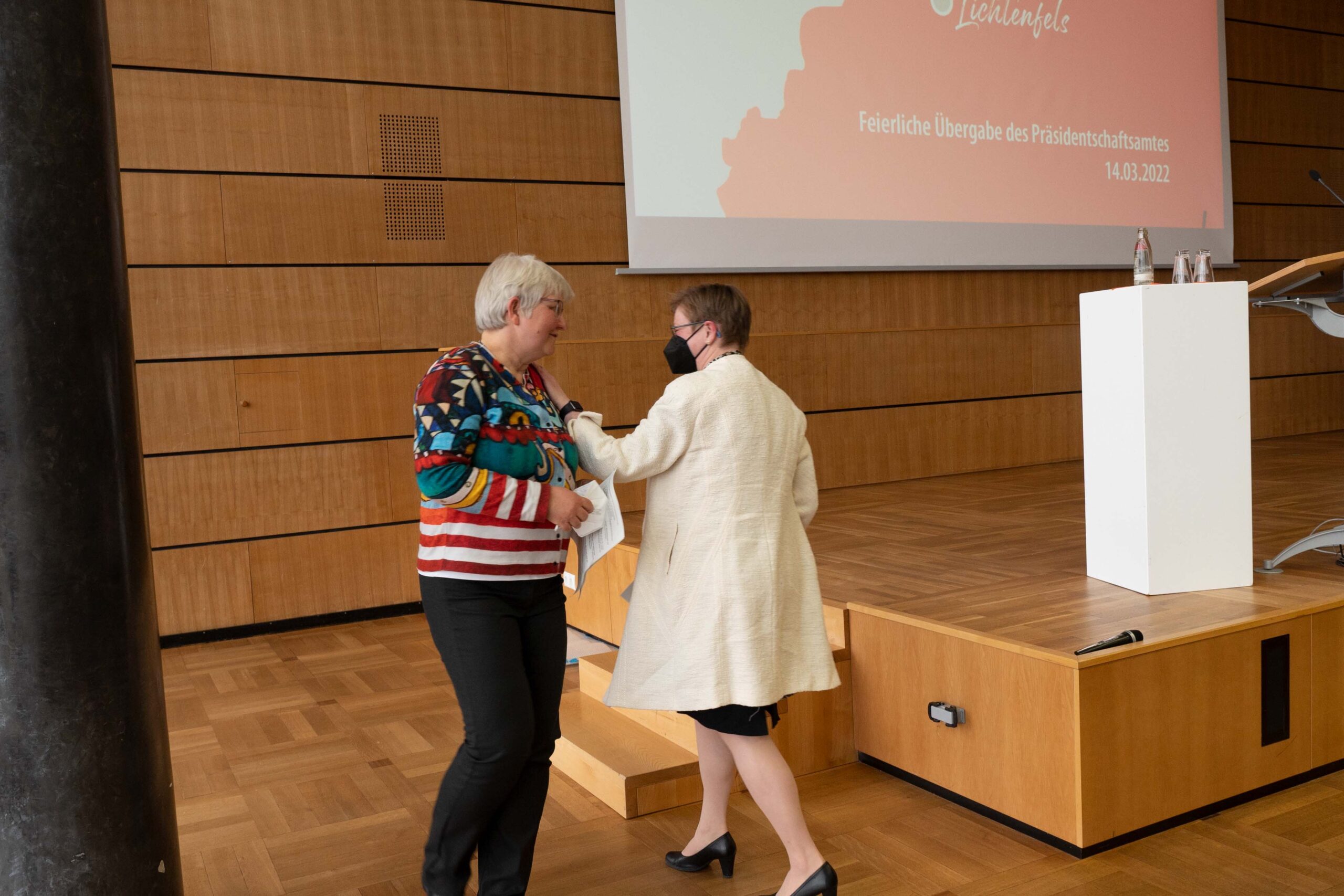 Zwei Frauen interagieren im Hörsaal der Hochschule Coburg, im Hintergrund eine Holzbühne mit großer Leinwand. Die eine trägt einen bunten Pullover, die andere einen weißen Kittel und eine Maske. Es scheint sich um eine feierliche Zeremonie am 14. Oktober 2022 zu handeln.