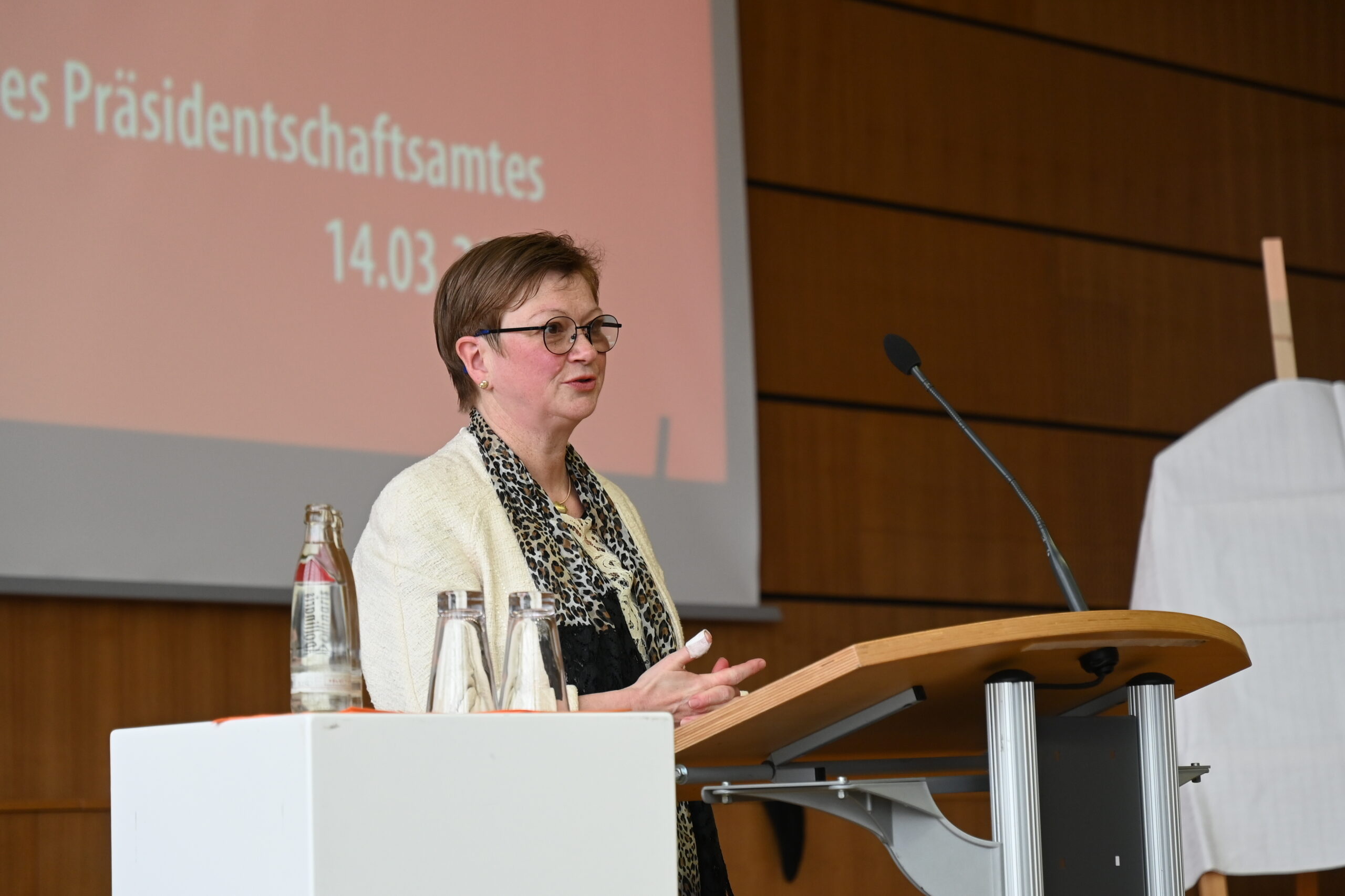 Eine Person mit kurzen Haaren und Brille steht an einem Podium, vermutlich an der Hochschule Coburg, und hält einen Vortrag. Sie trägt ein Kopftuch und eine helle Jacke. Hinter ihr ist ein großer Bildschirm zu sehen, auf dem ein deutscher Text mit dem Datum 14.03.23 angezeigt wird. Rechts ist ein verdecktes Objekt zu sehen.