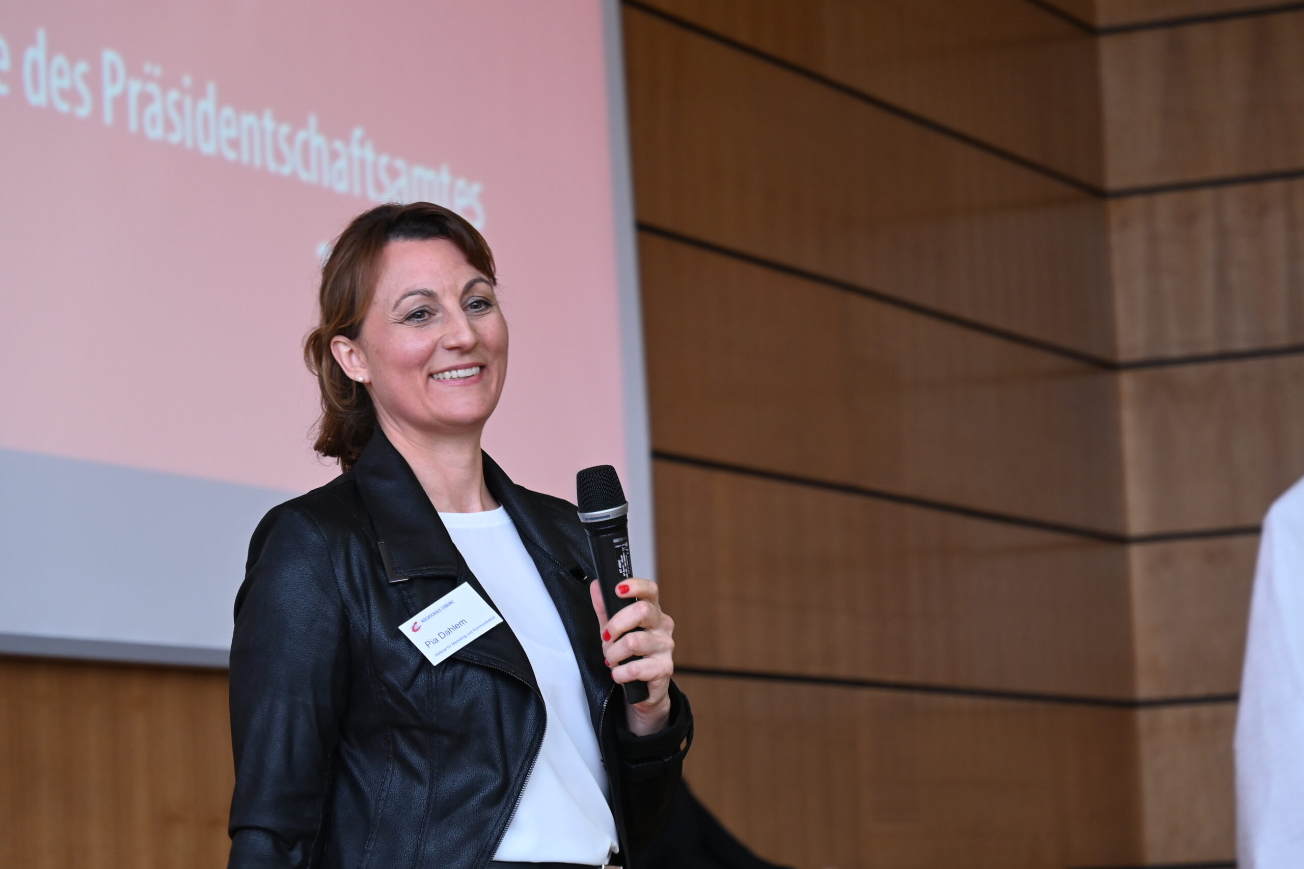 Eine Person mit einem Mikrofon steht lächelnd vor einer Holzwand. Im Hintergrund ist ein großer Bildschirm mit deutschen Texten über die Hochschule Coburg zu sehen. Die Person trägt eine schwarze Jacke und ein Konferenzabzeichen.