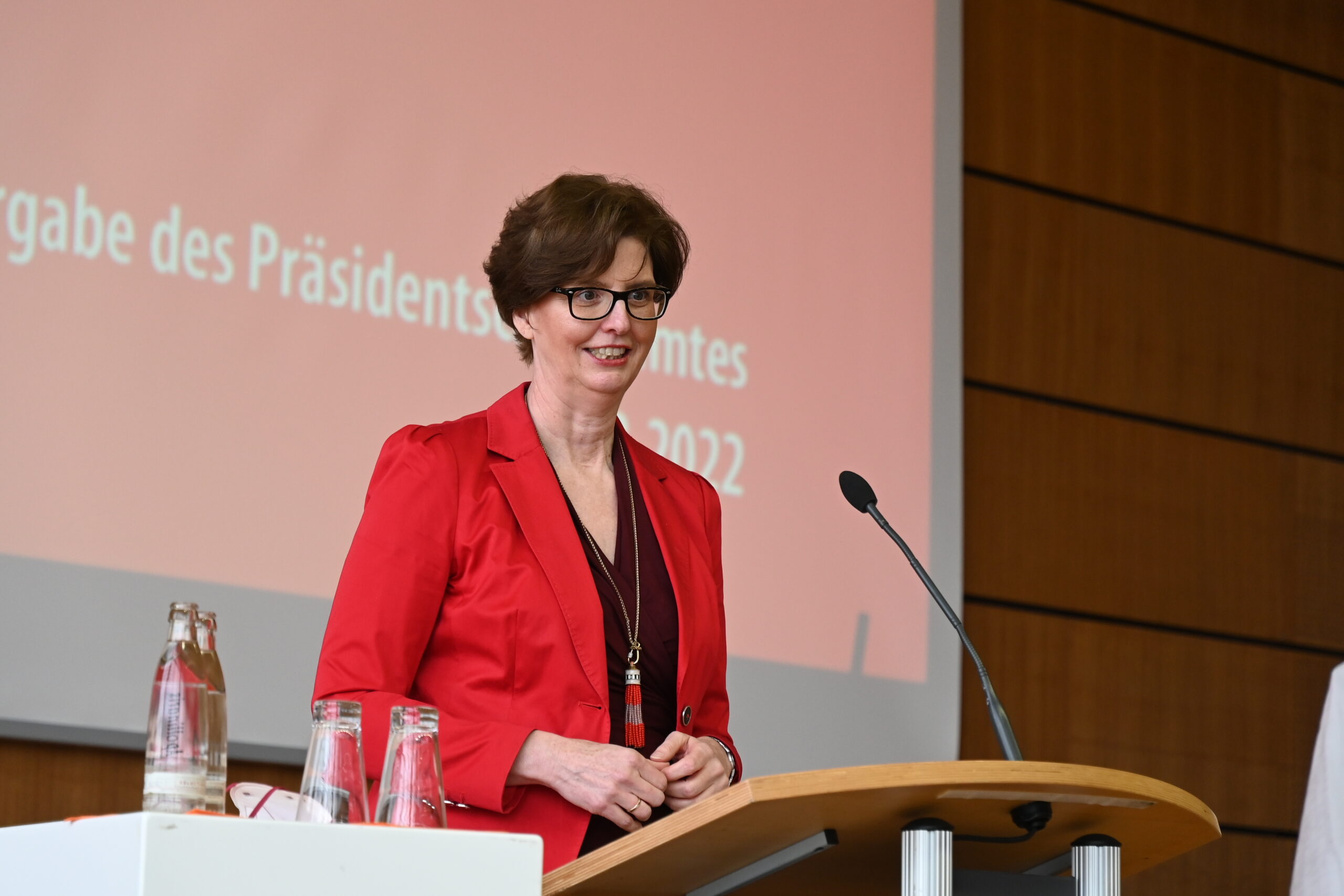 Eine Person in einer roten Jacke steht mit einem Mikrofon an einem Podium und spricht zu einem Publikum, das sich zu einer Veranstaltung an der Hochschule Coburg versammelt hat. Im Hintergrund ist ein Präsentationsbildschirm zu sehen, der die akademische Atmosphäre verstärkt.