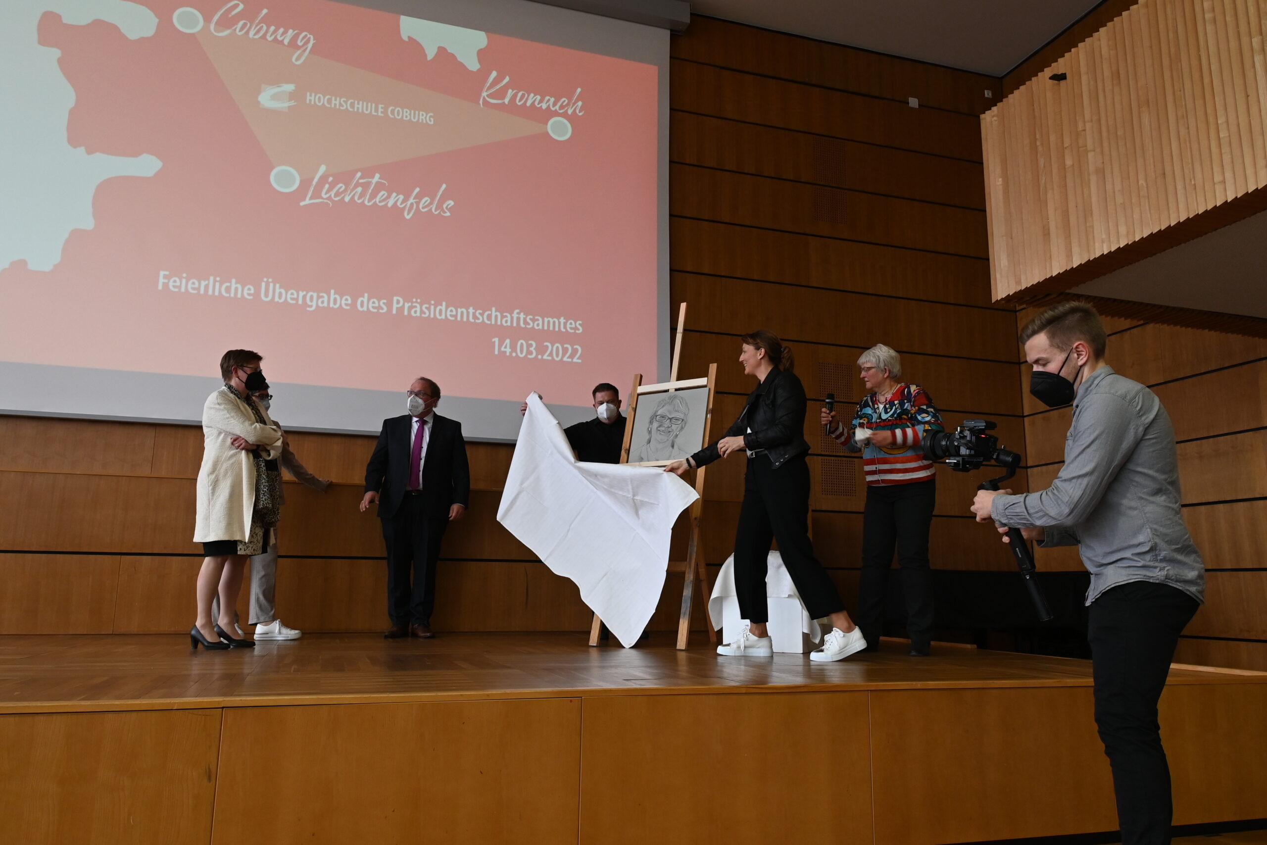 Während einer Zeremonie an der Hochschule Coburg steht eine Gruppe auf der Bühne, während einige einen gerahmten Gegenstand auspacken, während andere filmen. Auf dem Hintergrundbildschirm werden das Datum der Veranstaltung, 14.03.2022, und die Orte angezeigt: Coburg, Kronach, Lichtenfels.