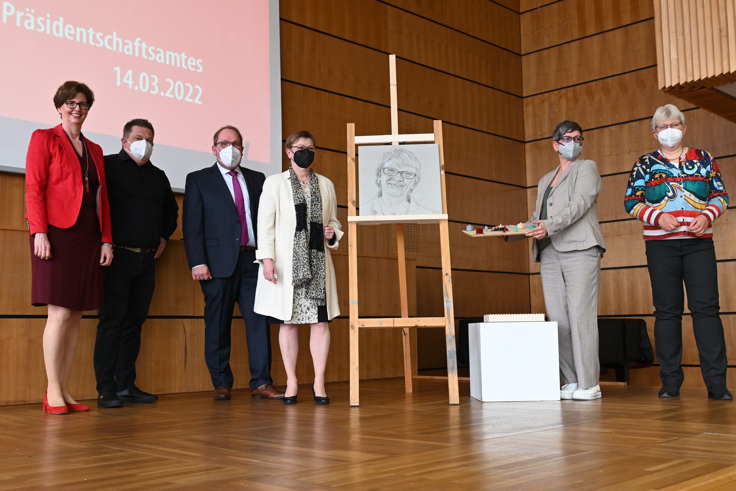 Eine Gruppe von sechs Personen mit Masken steht in einem Innenbereich neben einer Staffelei, auf der eine Skizze von einer von ihnen hängt. An der holzgetäfelten Wand im Hintergrund der Hochschule Coburg ist ein Bildschirm mit dem Datum 14.03.2022 zu sehen.