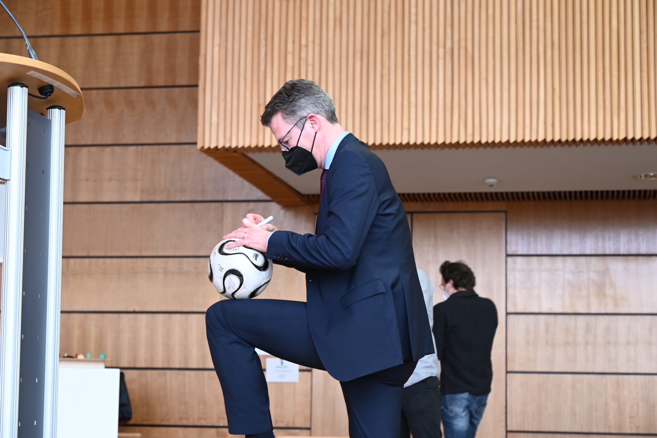 Ein Mann in Anzug und Gesichtsmaske steht in einem Innenbereich der Hochschule Coburg und signiert einen Fußball, während er einen Fuß auf einer Stufe abstellt. Den Hintergrund bildet eine Holzvertäfelung, im Hintergrund ist eine weitere Person zu sehen, die weggeht.