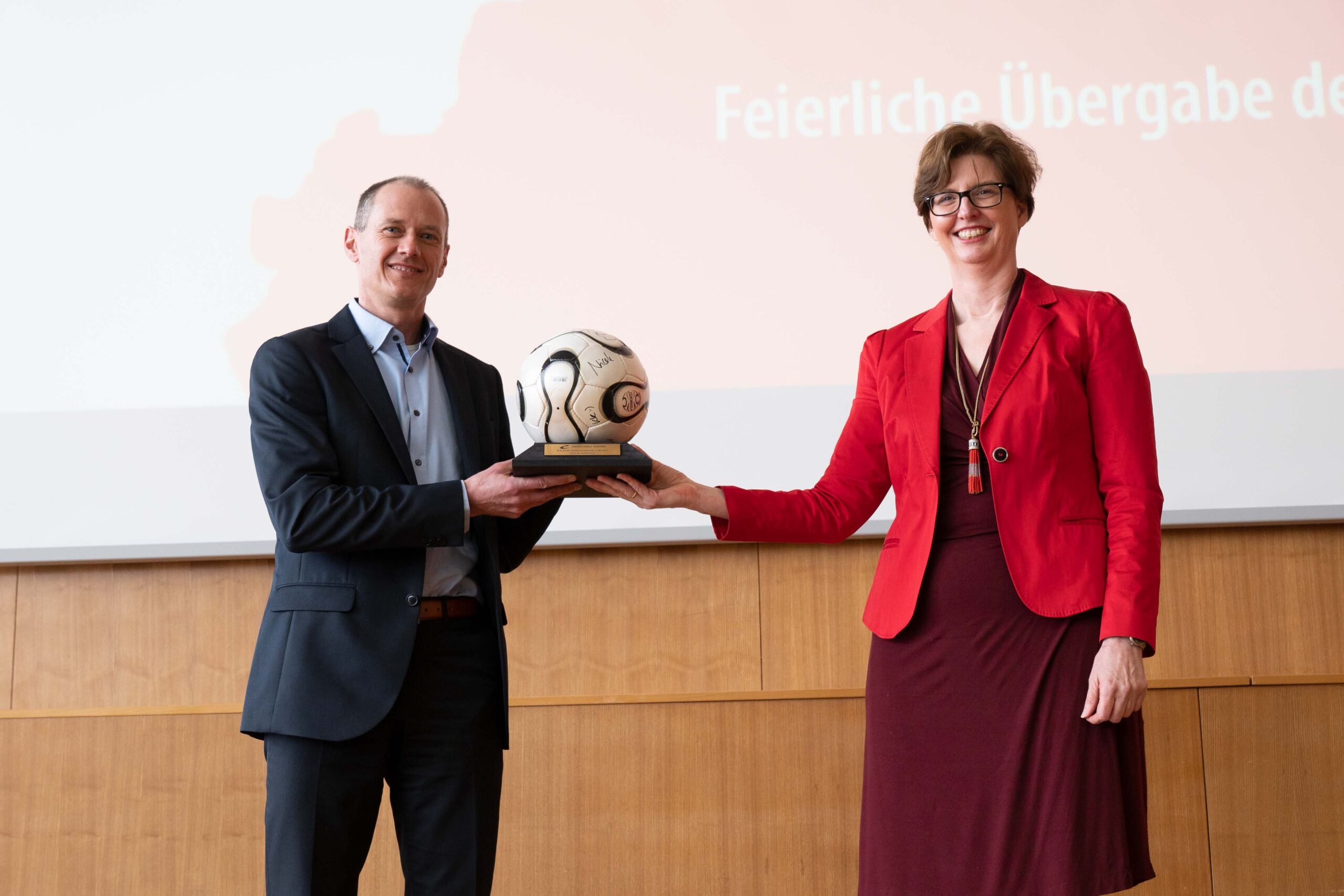 Zwei Menschen stehen nebeneinander und halten jeweils einen Pokal in der Hand, der einen Fußball auf einem Holzsockel zeigt. Der Mann links trägt einen Anzug, die Frau rechts ein rotes Outfit. Hinter ihnen zeigt eine Projektionsleinwand einen Text auf Deutsch, der auf eine Veranstaltung an der Hochschule Coburg aufmerksam macht.