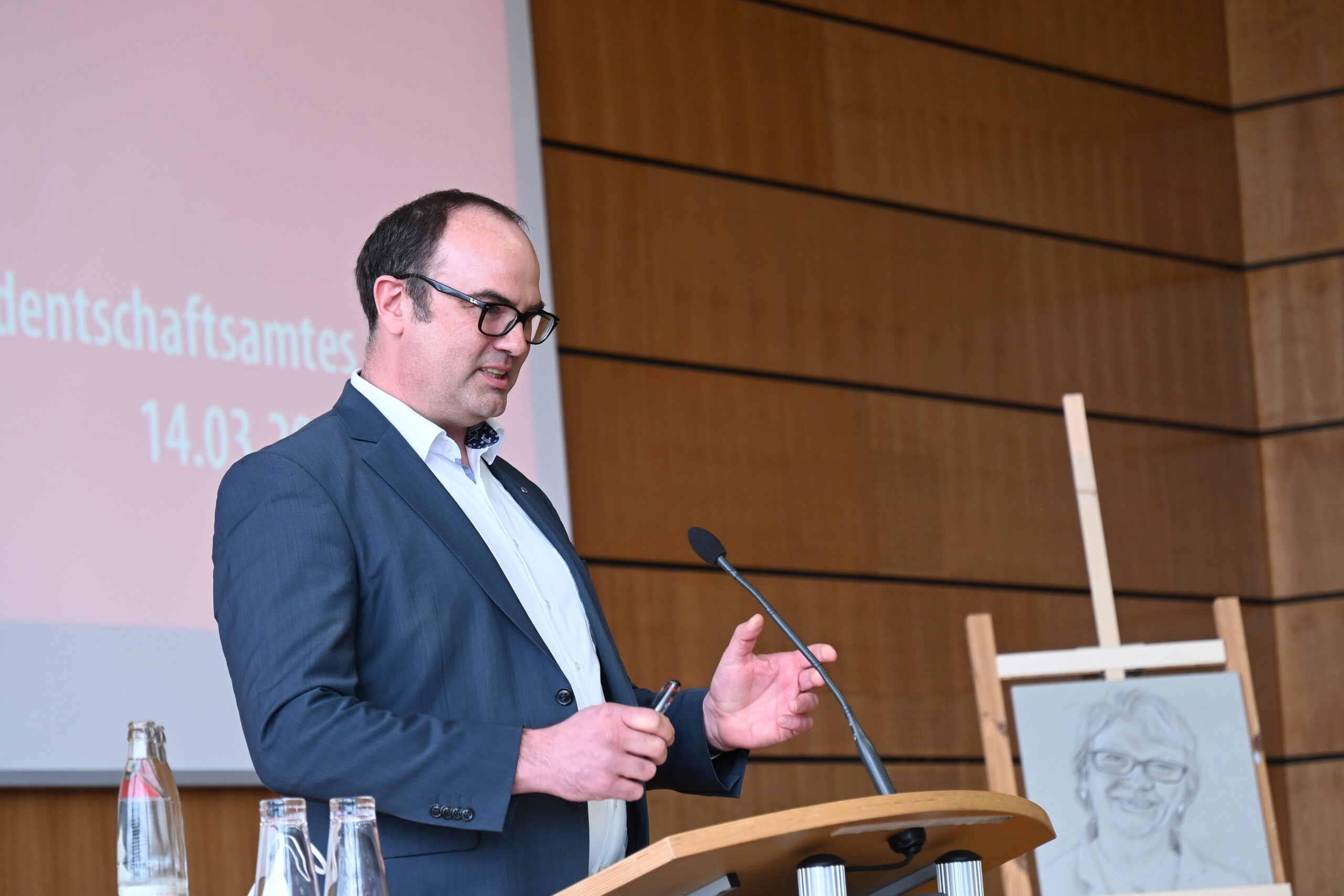 Ein Mann in Anzug und Brille spricht an einem Podium in einem Konferenzraum der Hochschule Coburg. Hinter ihm stehen ein Mikrofon und ein Porträt auf einer Staffelei. Auf dem Tisch daneben stehen eine Flasche und ein Glas, im Hintergrund eine Holzpaneelwand.