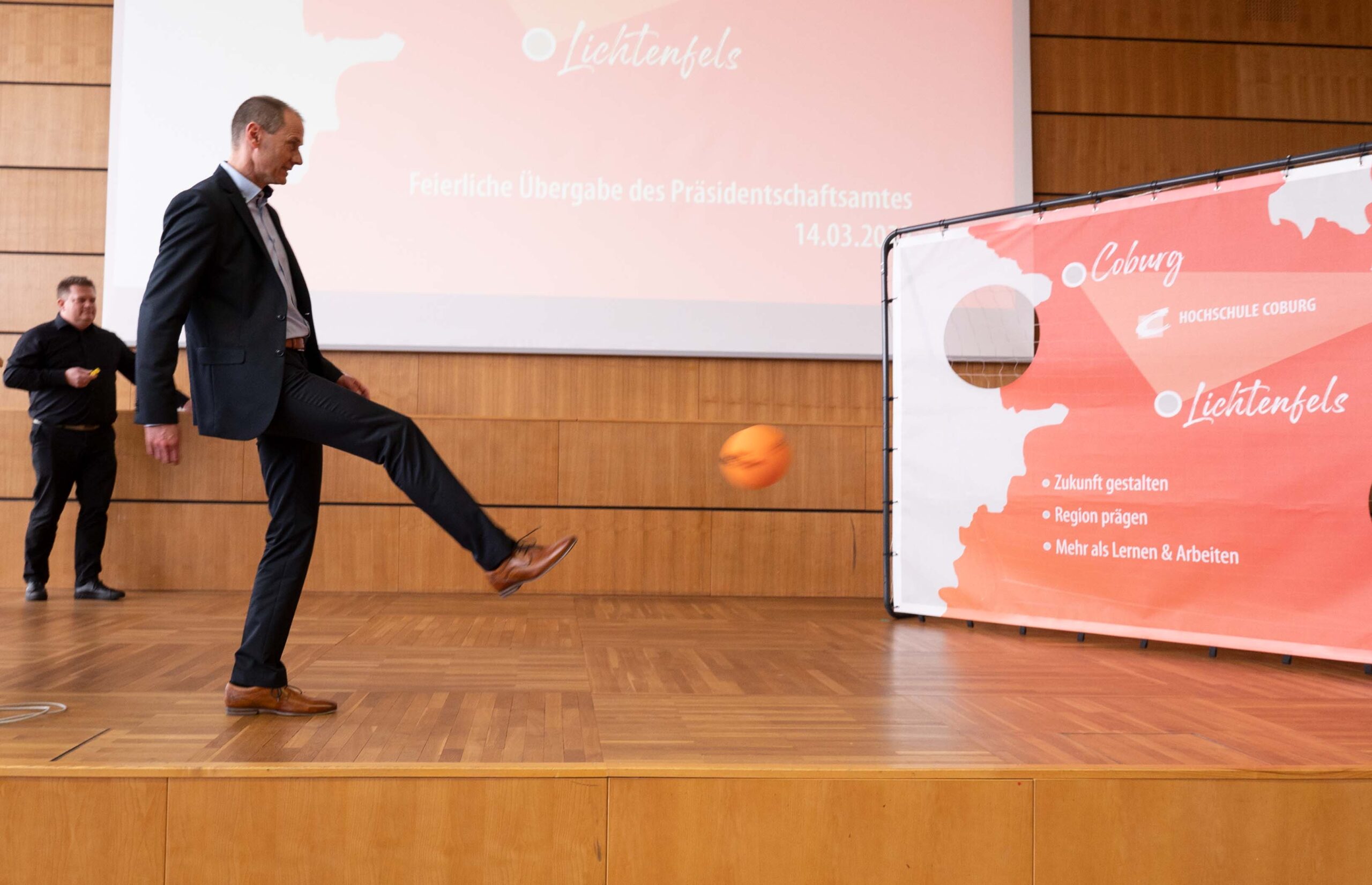 Ein Mann im Anzug kickt einen orangefarbenen Ball in der Nähe eines großen Bildschirms, auf dem Text und das Logo der Hochschule Coburg zu sehen sind. Ein weiterer Mann steht im Hintergrund. Es scheint sich um eine formelle Veranstaltung oder Präsentation zu handeln.