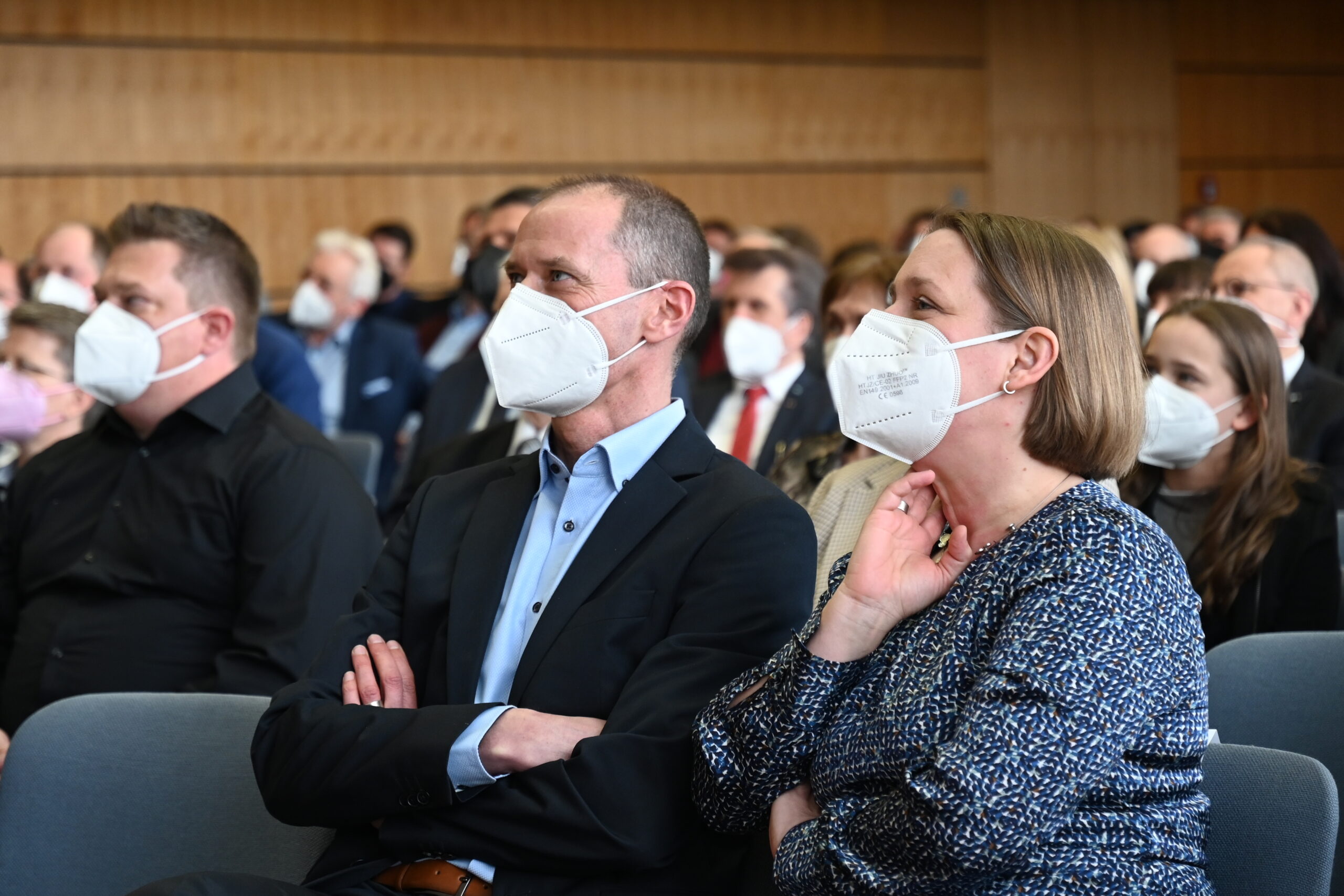 Eine Gruppe von Menschen sitzt in einem Hörsaal der Hochschule Coburg, alle tragen weiße Mund-Nasen-Masken. Sie wirken aufmerksam, sind formell gekleidet und sitzen eng beieinander. Die Atmosphäre lässt auf eine Konferenz oder ein Seminar schließen.