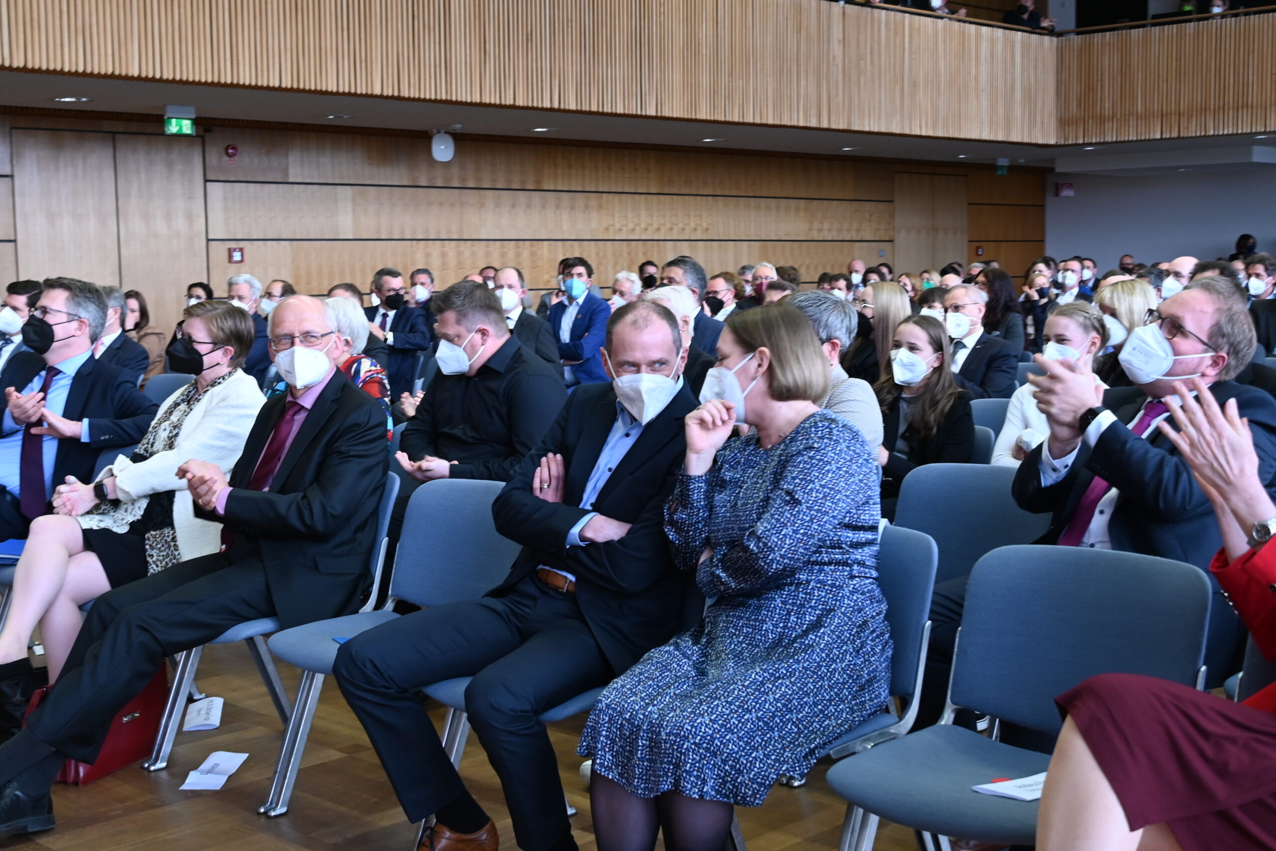 Eine große Gruppe von Menschen sitzt in Stuhlreihen in einem Konferenzsaal der Hochschule Coburg. Viele tragen Gesichtsmasken und sind in formelle Kleidung gekleidet. Sie wirken engagiert, einige klatschen. Die Umgebung strahlt eine professionelle oder akademische Atmosphäre aus.